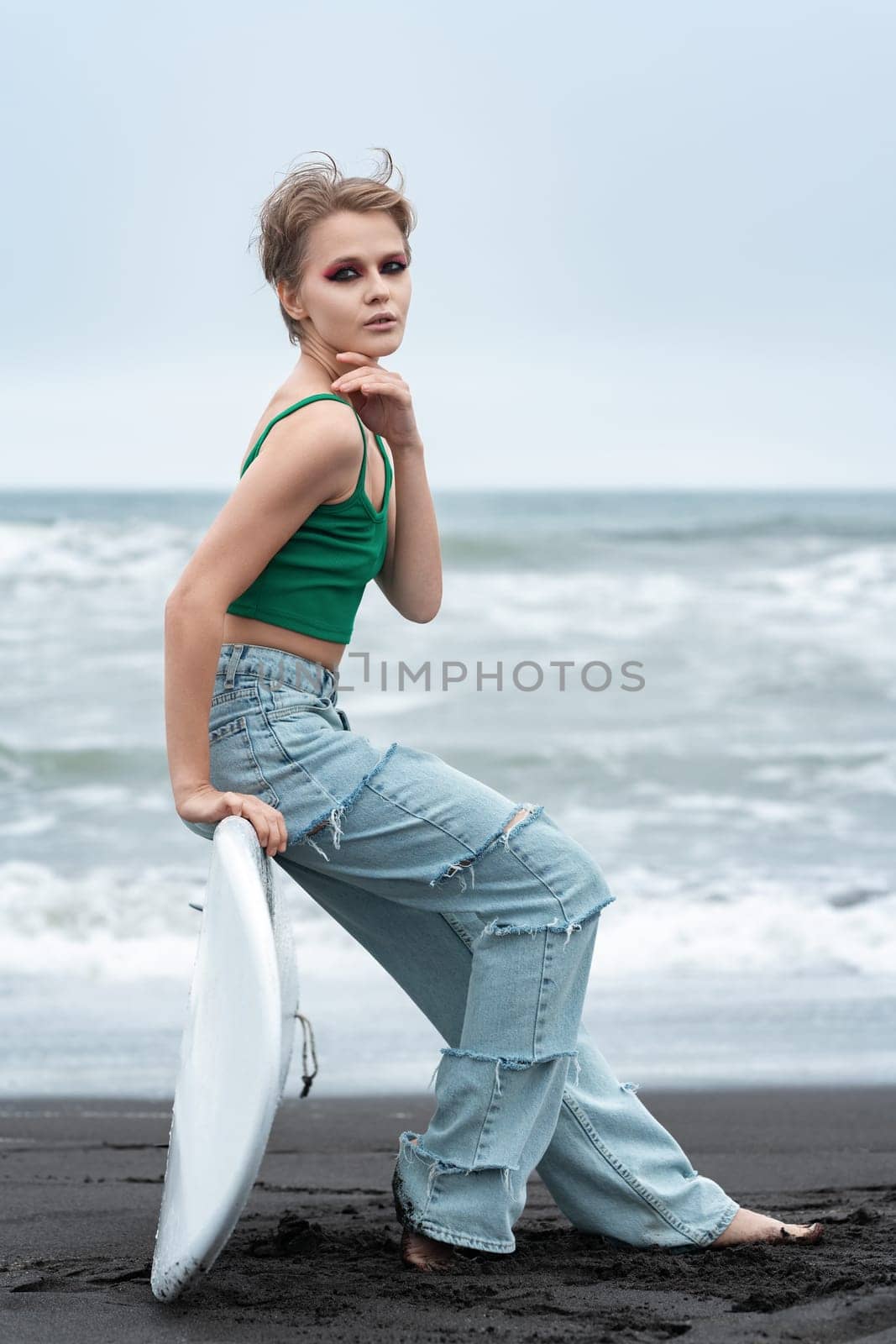 Portrait hipster woman surfer sitting on surf board lying on sand, on background of sea waves. Side view of Caucasian model with short hair and bright makeup looking at camera, wearing top and jeans