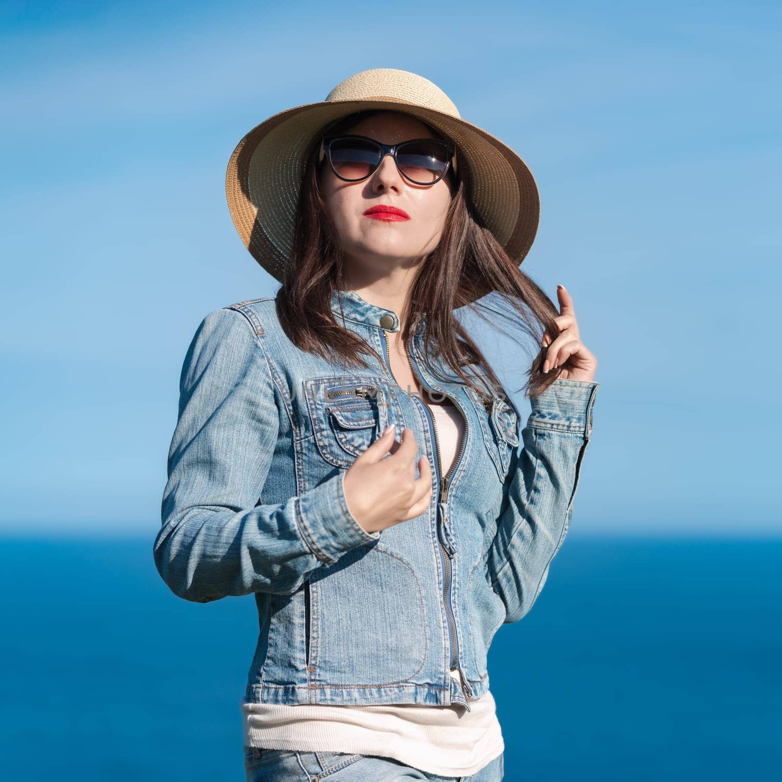 Portrait of brunette female in denim jacket, jeans, straw hat on head and sunglasses looking at camera. Hipster adult woman standing on background of blue sky and ocean on summer sunny day