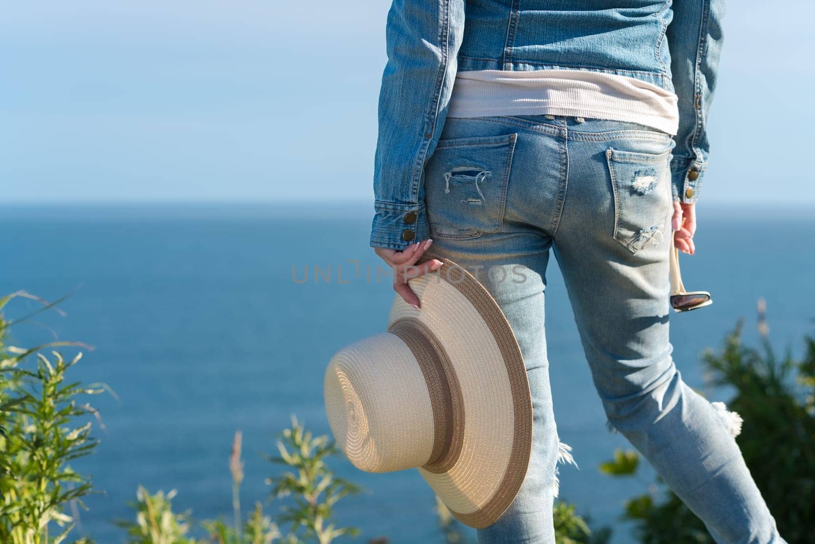 Cropped rear view below waist on legs of woman in blue jeans. Unrecognizable hipster female holding straw hat in one hand and sunglasses in other, standing in grass on seashore in sunny summer weather