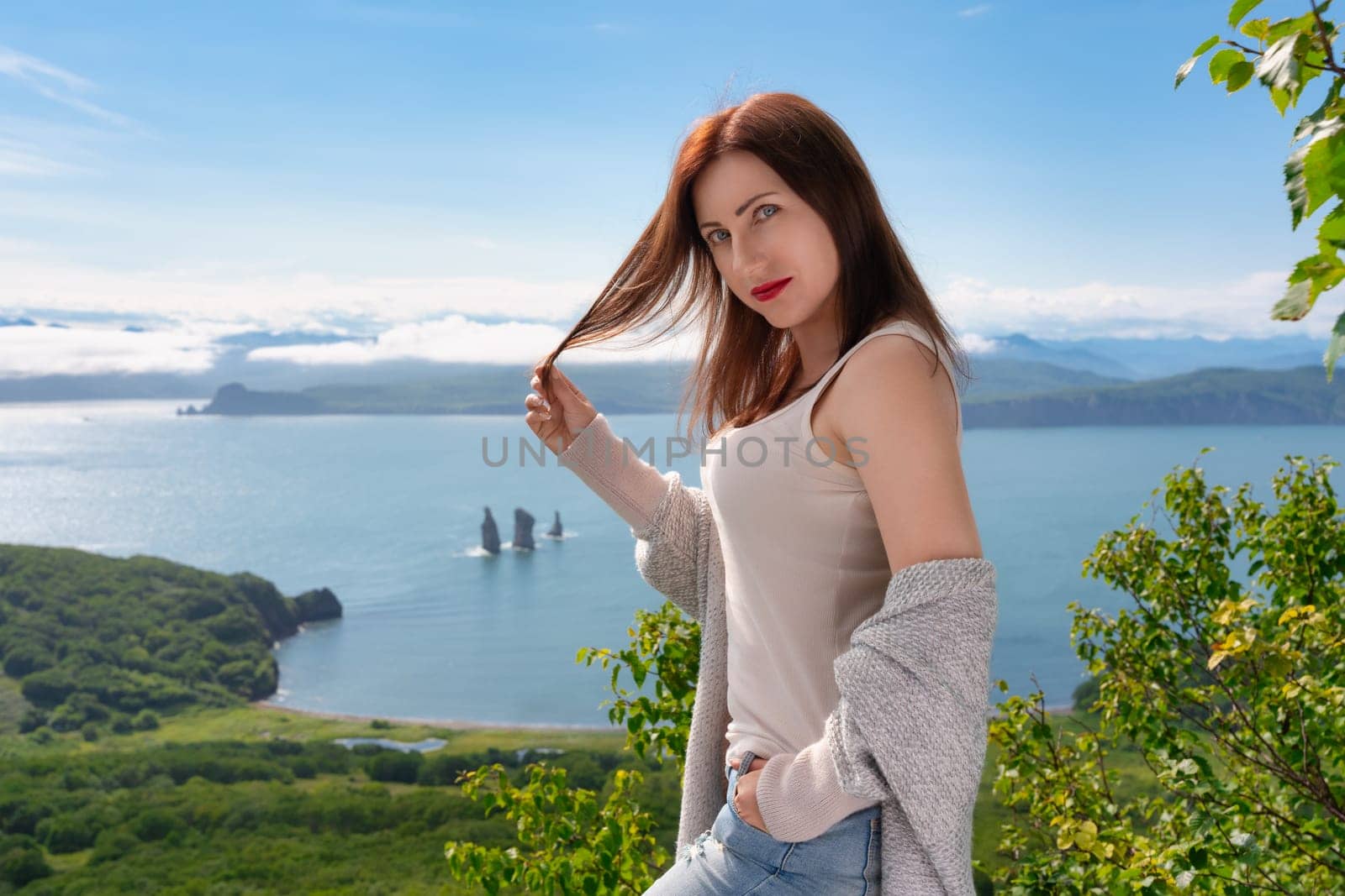Adult Caucasian ethnicity woman in light T-shirt standing on high mountain shore against panoramic view of sea on sunny summer day. Brunette woman looking at camera