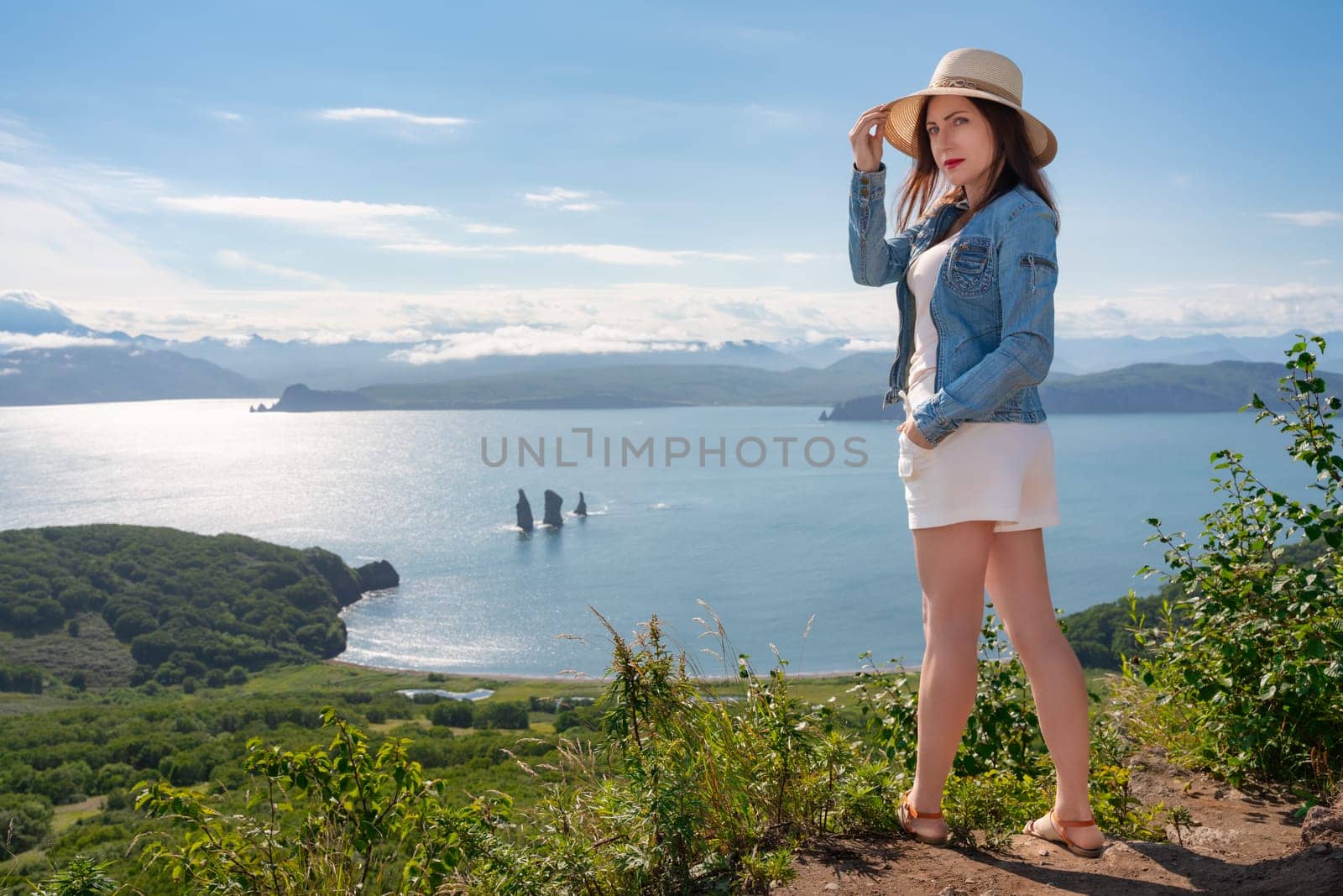 Hipster woman looking at camera, dressed in straw hat, denim jacket, white shorts and sandals. Female tourist standing on high mountain against backdrop of panoramic ocean view on sunny summer day