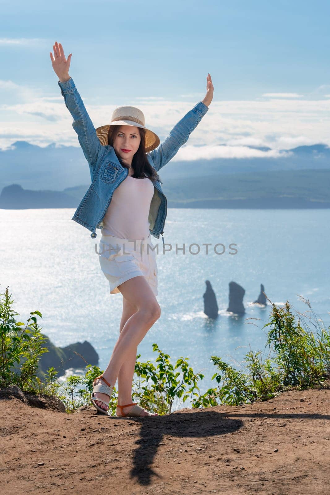Hipster woman looking at camera with her hands raised, dressed in straw hat, denim jacket, white shorts and sandals, stands on high mountain against background of ocean on sunny summer day