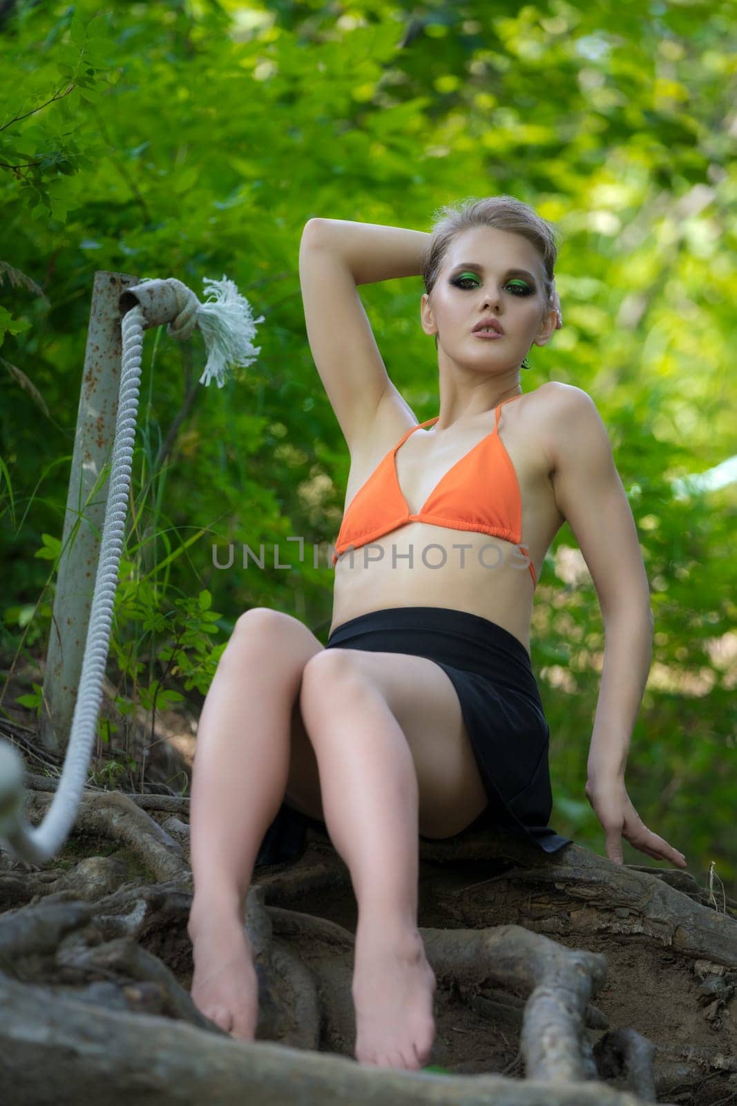 Sensuality woman sitting and poses in forest on ladder made of tree roots. Caucasian adult female dressed in orange bikini top, mini skirt. Blonde model with short hair and make-up, looking at camera