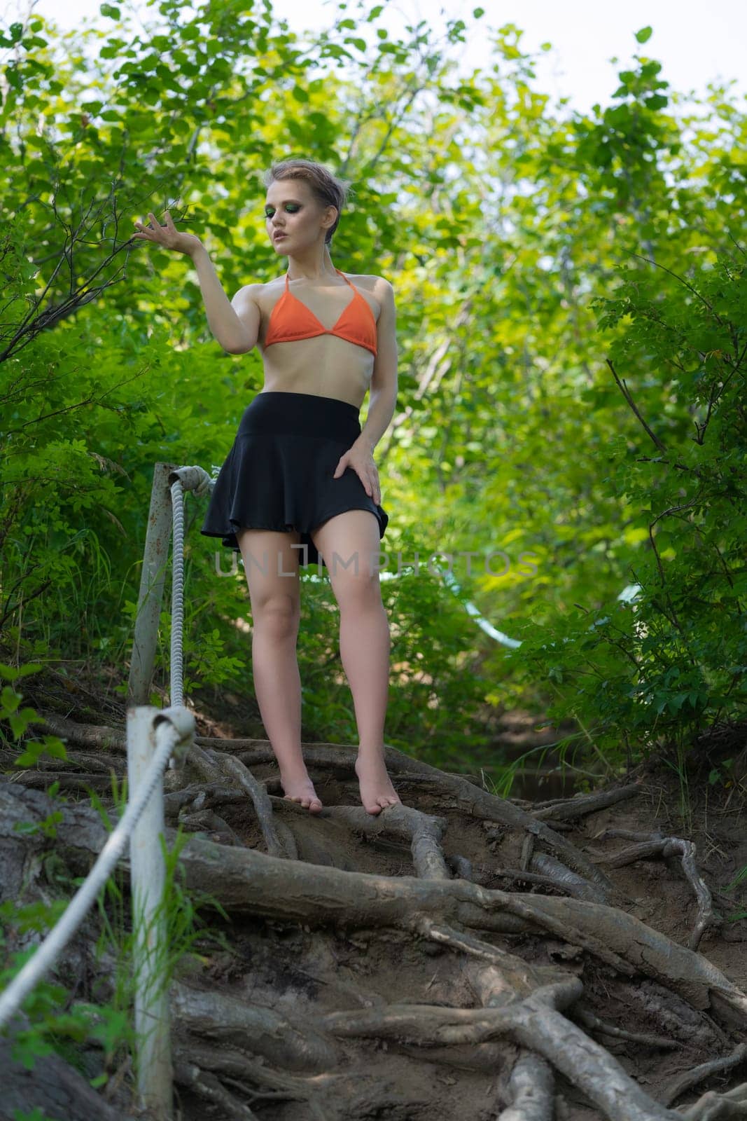 Caucasian woman standing and poses in forest on ladder made of tree roots. Sensuality adult female dressed in orange bikini top and mini skirt. Blonde model with short hair and bright make-up
