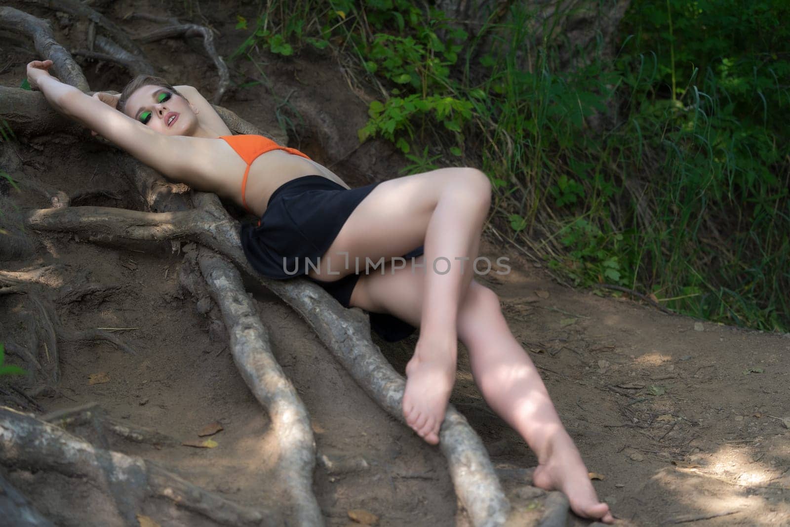 Sensual slim woman model with long legs lying down on ground on tree roots in forest. Sexual adult woman dressed in bikini top and mini skirt. Playful blonde with short hair and make-up looking down