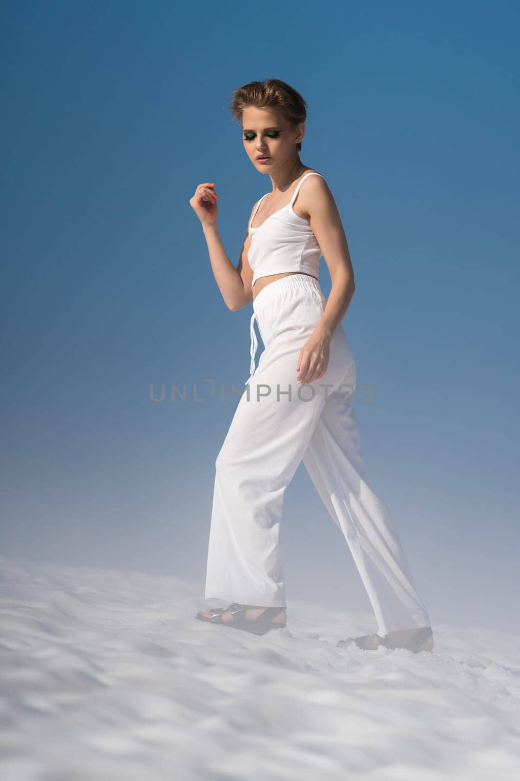 Beauty slim adult woman dressed in white crop top, white pants and sandals. European female tourist walking through snow with light fog on sunny weather with blue sky. Full length, side view