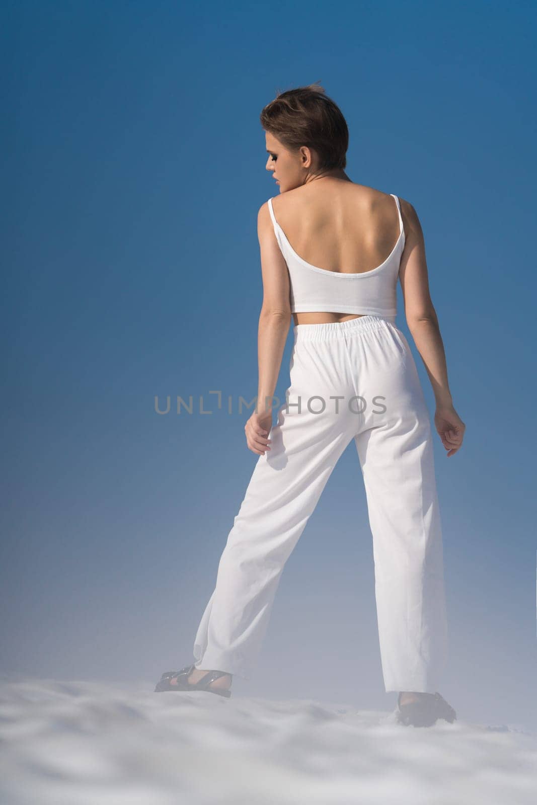 Rear view of fashionable woman posing through snow with light fog on sunny day with blue sky. Playful blond fashion model wearing in white cropped top and white pants, sandals. Full length