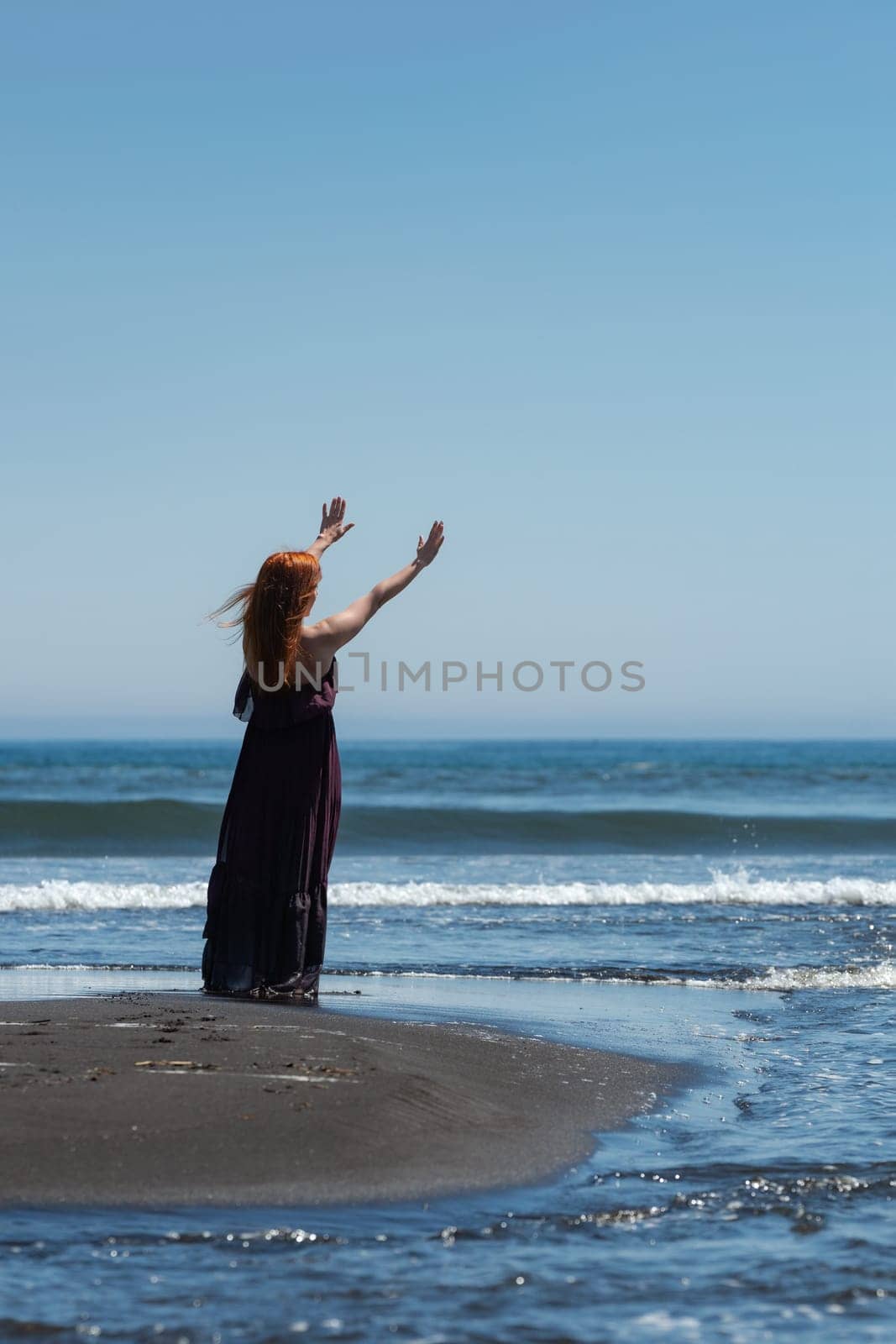 Woman in purple dress looks so happy and carefree as stands on sandy beach with arms raised high towards gorgeous blue sky and sea waves. She's even barefoot, which adds to feeling of freedom and joy