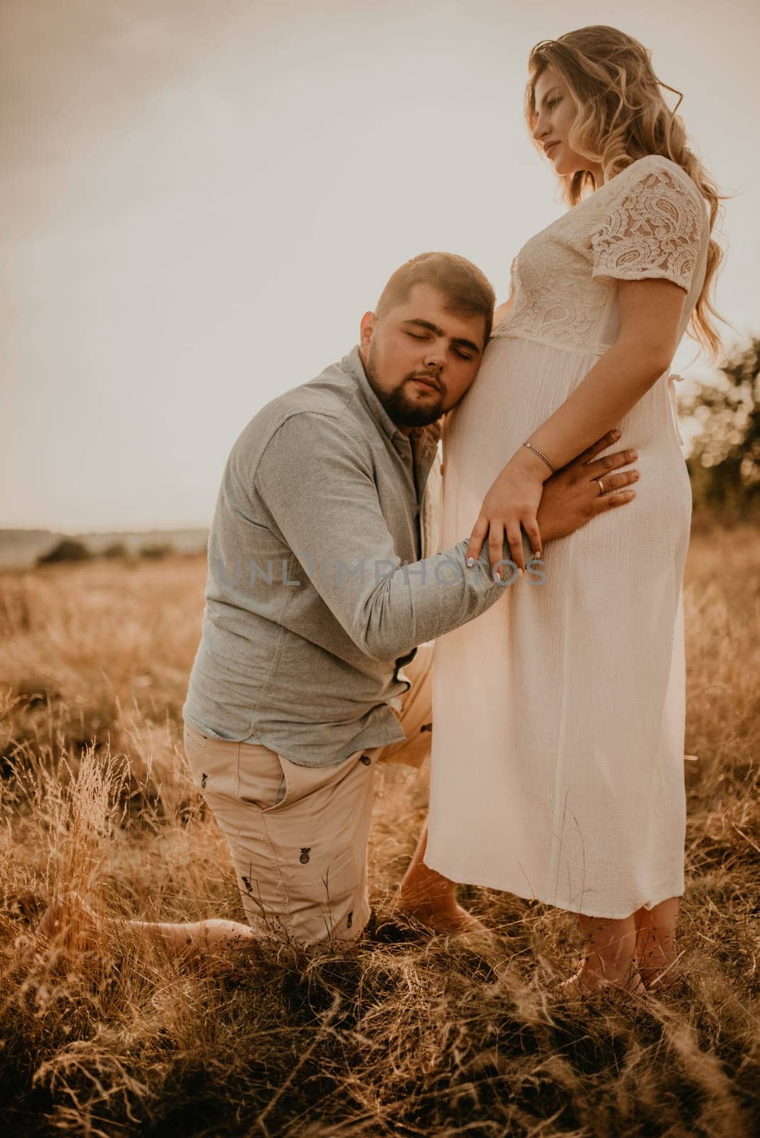 husband hugs pregnant wife father listens belly put his ear to tummy. Happy family resting in nature hugs kisses in summer at sunset. future mother Caucasian woman in white cotton dress