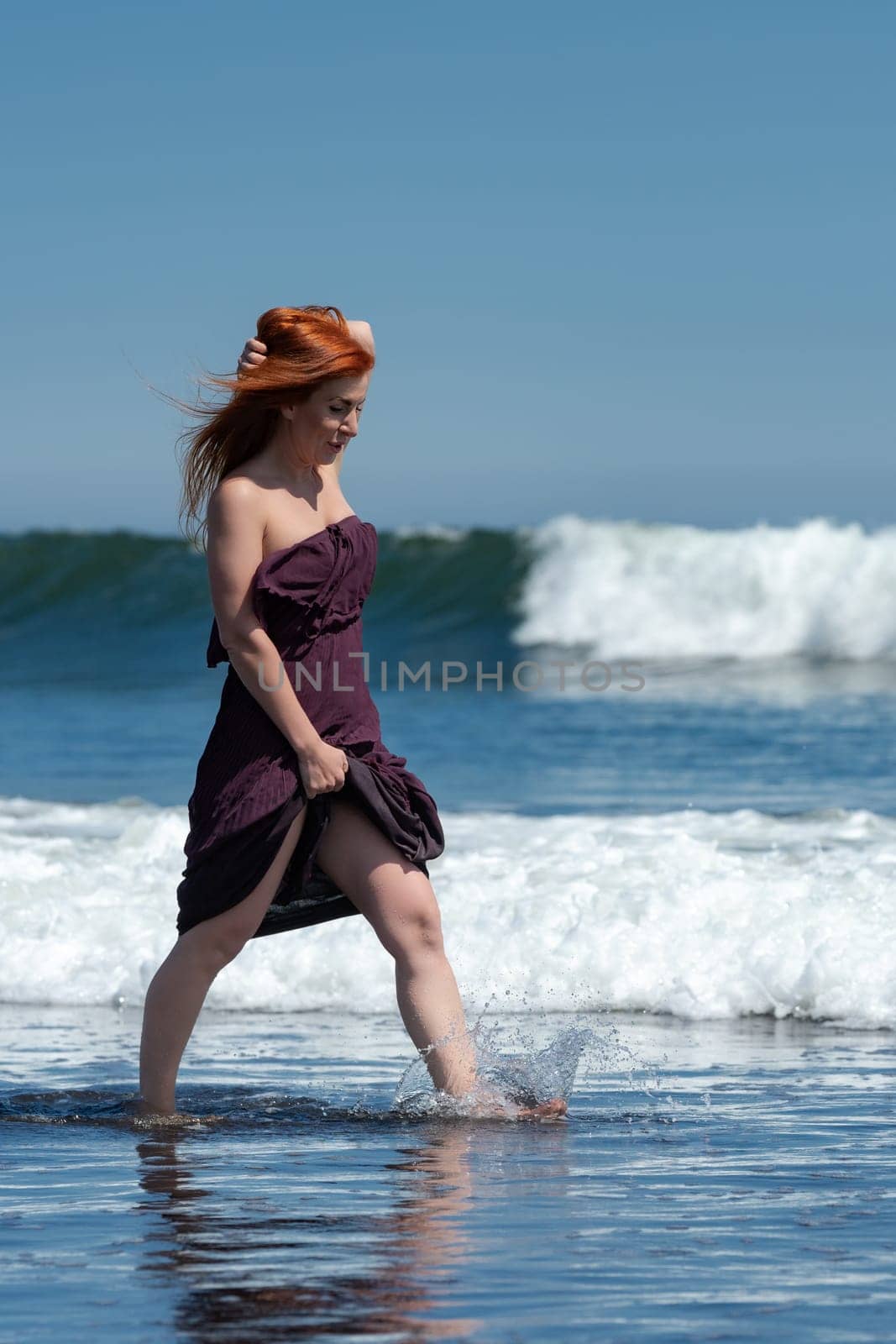 Happy redhead woman enjoying summer holidays walking ankle deep in water of breaking ocean waves on beach on sunny day. She looks stunning in dark puce long dress with knee high hem, moving barefoot