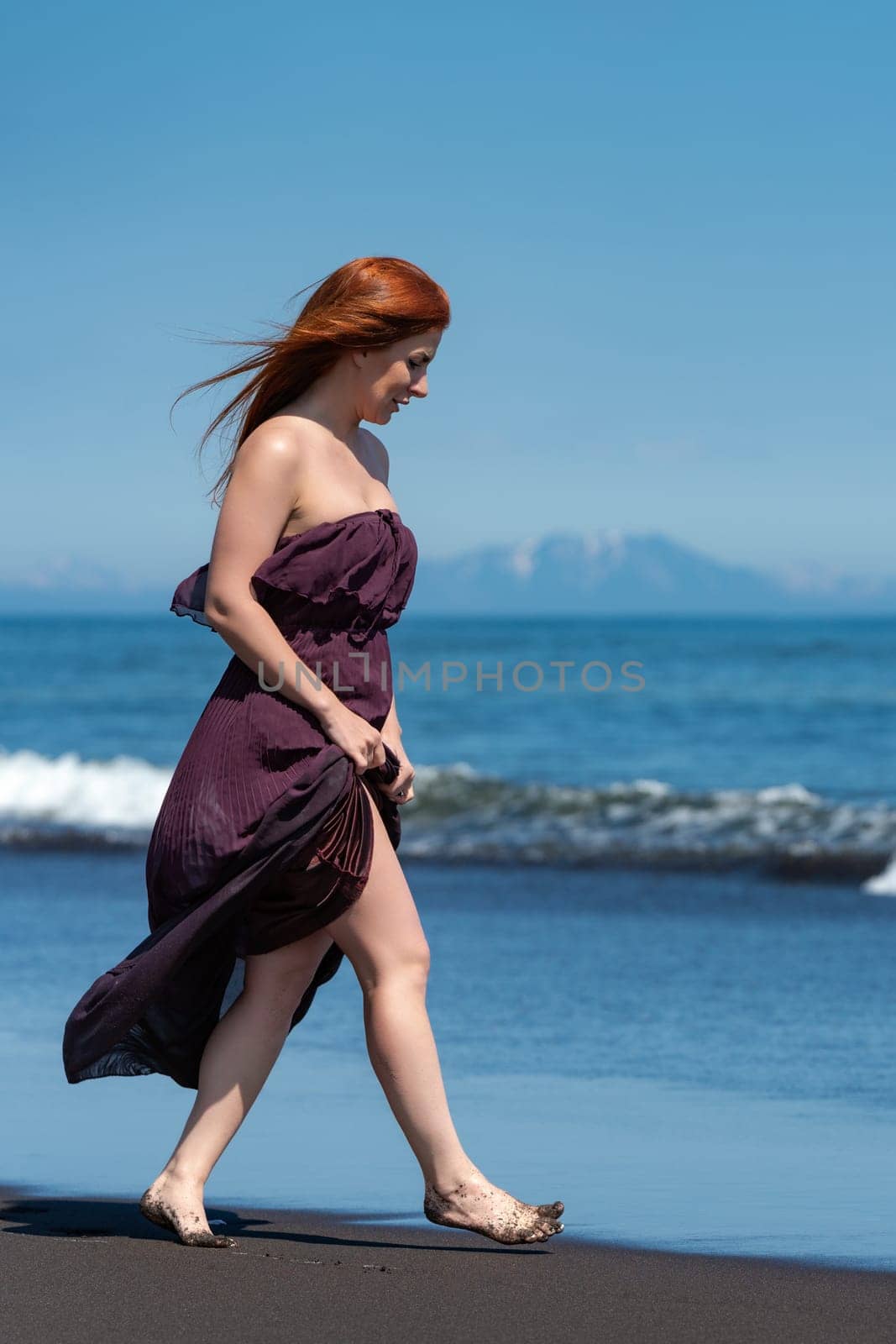 Redhead woman is walking along sandy beach in long dress with hem raised to knees. She looks down and enjoys summer holiday on ocean shore on sunny summer day. Full-length side view of barefoot female