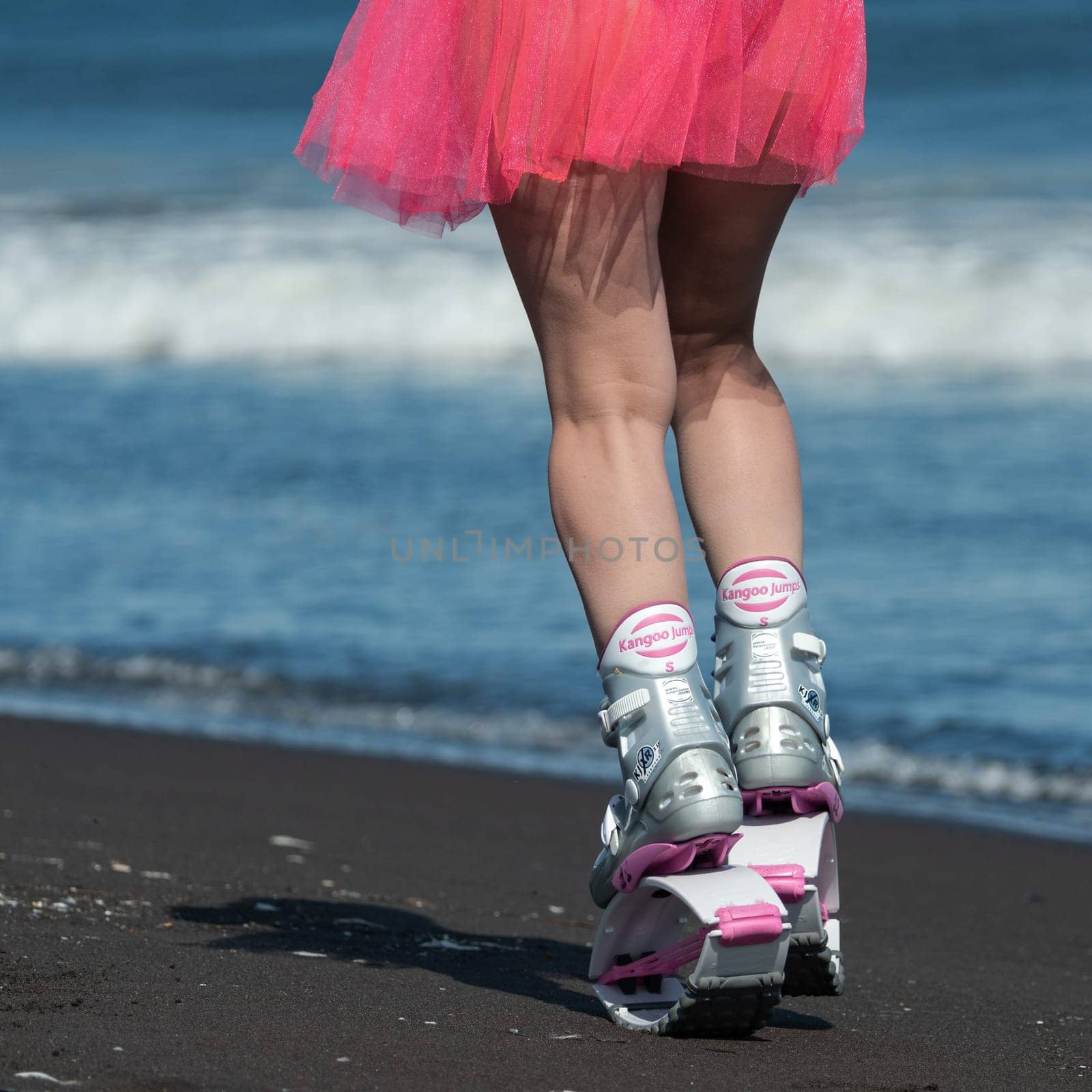 Rear view sportive woman legs in Kangoo Jumps sports boots and short pink skirt running on beach during aerobic exercise outdoors by Alexander-Piragis
