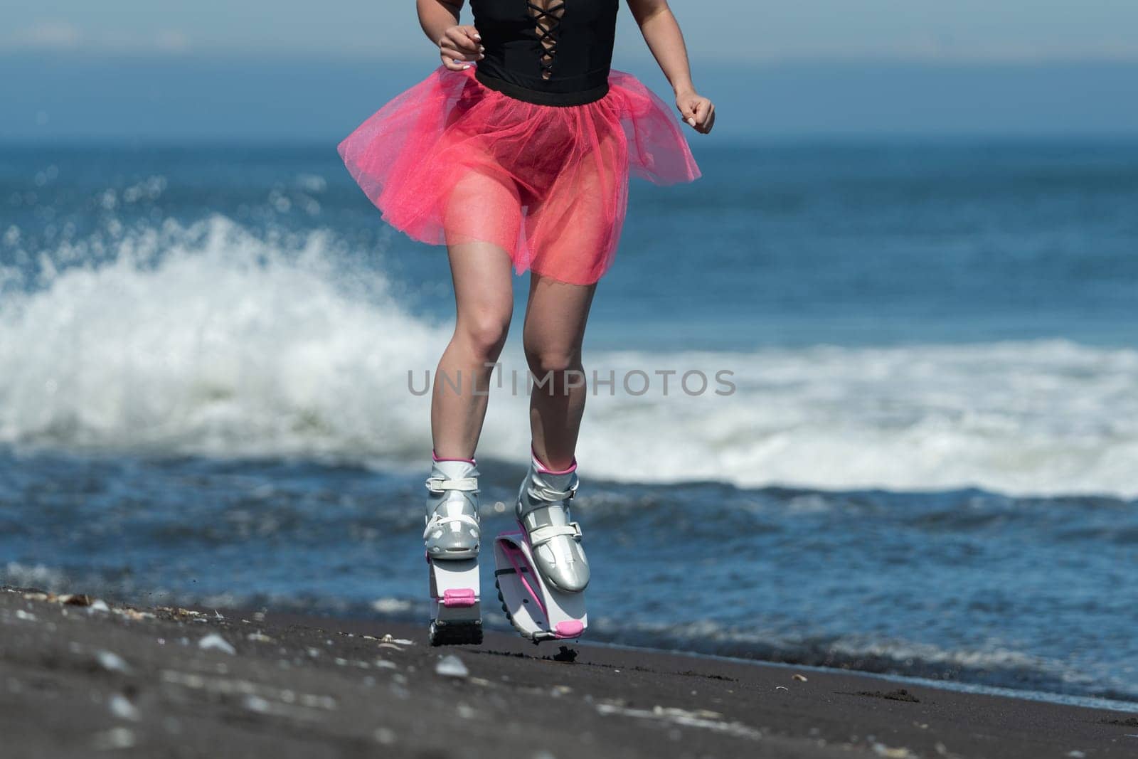 Sportswoman in sports Kangoo Jumps boots, swimsuit and short skirt running and jumping on sandy beach during aerobic session workout by Alexander-Piragis