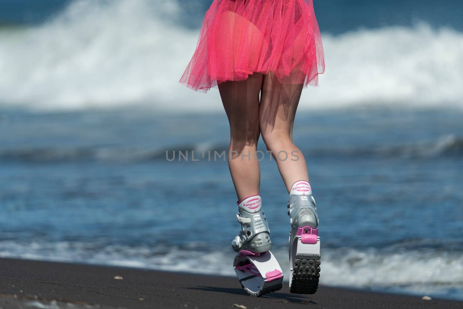 Rear view of fit female legs in short skirt and sports jumping boots Kangoo Jumps running on beach during training fitness session. Low section, crop view by Alexander-Piragis