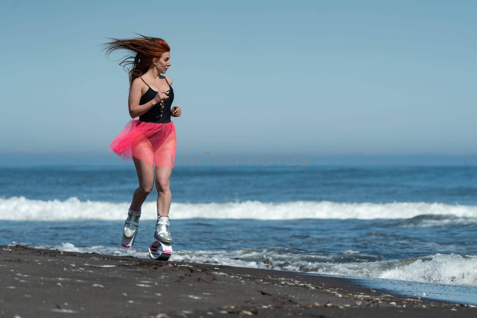 KAMCHATKA, RUSSIA - JUNE 15, 2022: Athletic woman in sports Kangoo Jumps boots, black one piece swimsuit, short skirt running and jumping on sandy beach during fitness and aerobic exercising outdoors