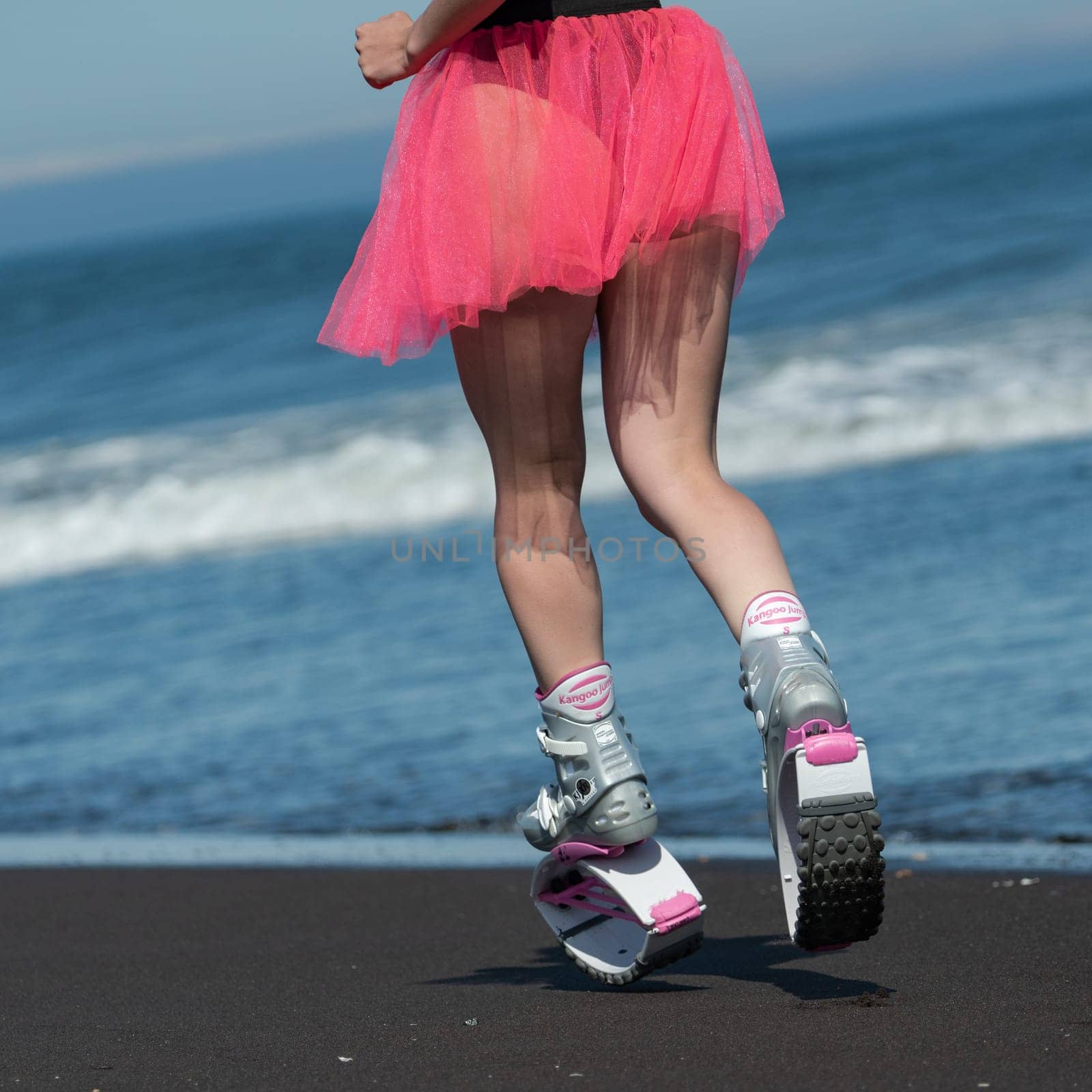 KAMCHATKA, RUSSIA - JUNE 15, 2022: Rear view of female legs in sports Kangoo Jumps boots, swimsuit and short pink skirt running and jumping on sandy beach during fitness training. Dutch angle shot