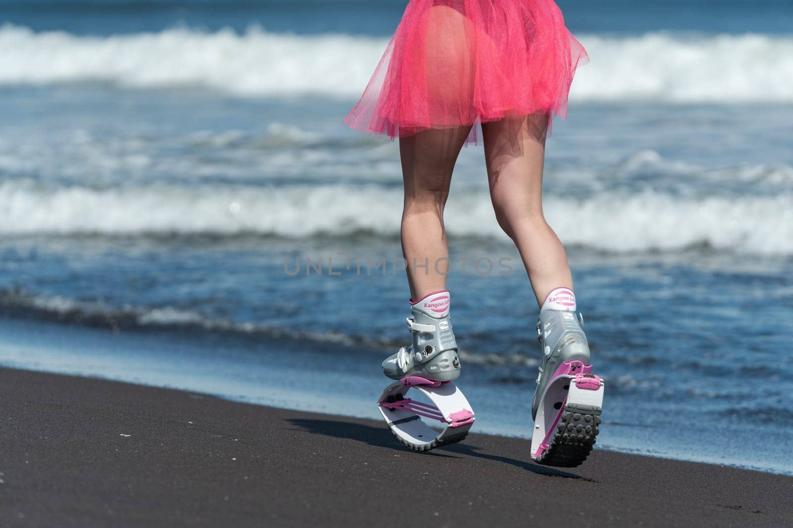 Rear view woman running and jumping on beach during aerobic workout. Female in Kangoo Jumps boots and short pink skirt. Low section, crop view by Alexander-Piragis