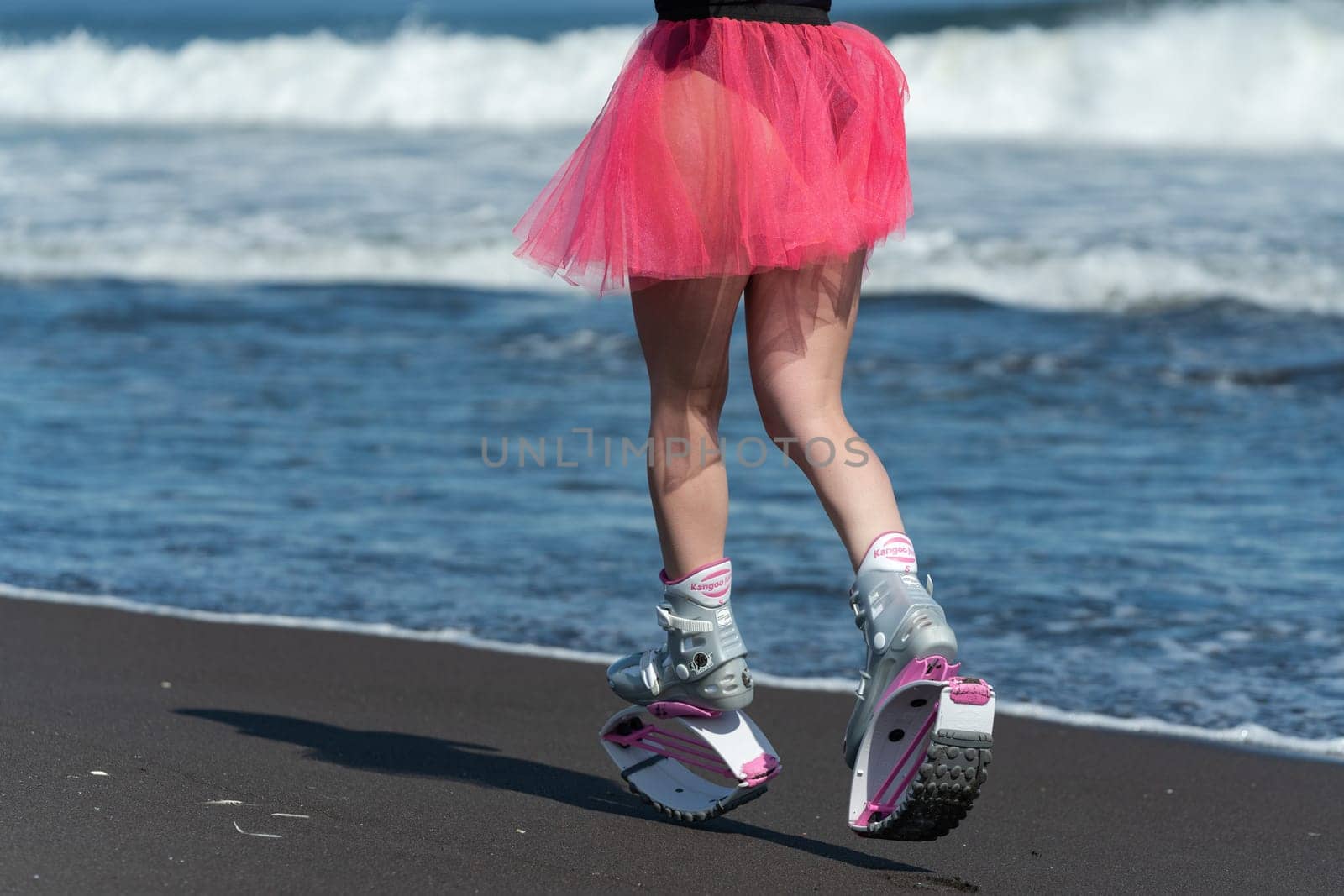 KAMCHATKA, RUSSIA - JUNE 15, 2022: Rear view of woman legs in sports boots Kangoo Jumps, black swimsuit and short skirt running on black sandy beach. Outdoors fitness workout on summer sea shore