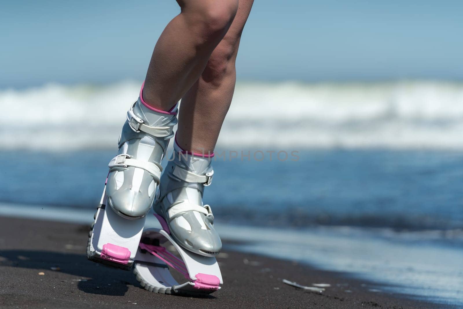 KAMCHATKA, RUSSIA - JUNE 15, 2022: Female legs in sports Kangoo Jumps boots jumping and running on black sandy beach during fitness training session outdoors on seashore on background of ocean waves