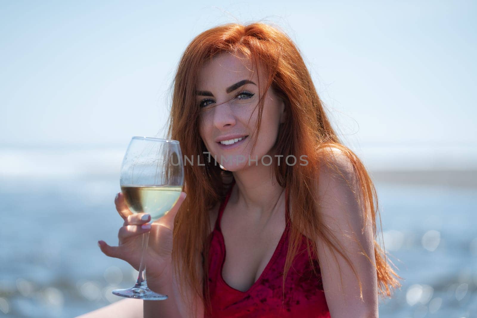 Portrait of smiling redhead woman holding wineglass and looking at camera against bokeh background of ocean waves at beach party celebration during summer holidays. Happy woman in red evening dress