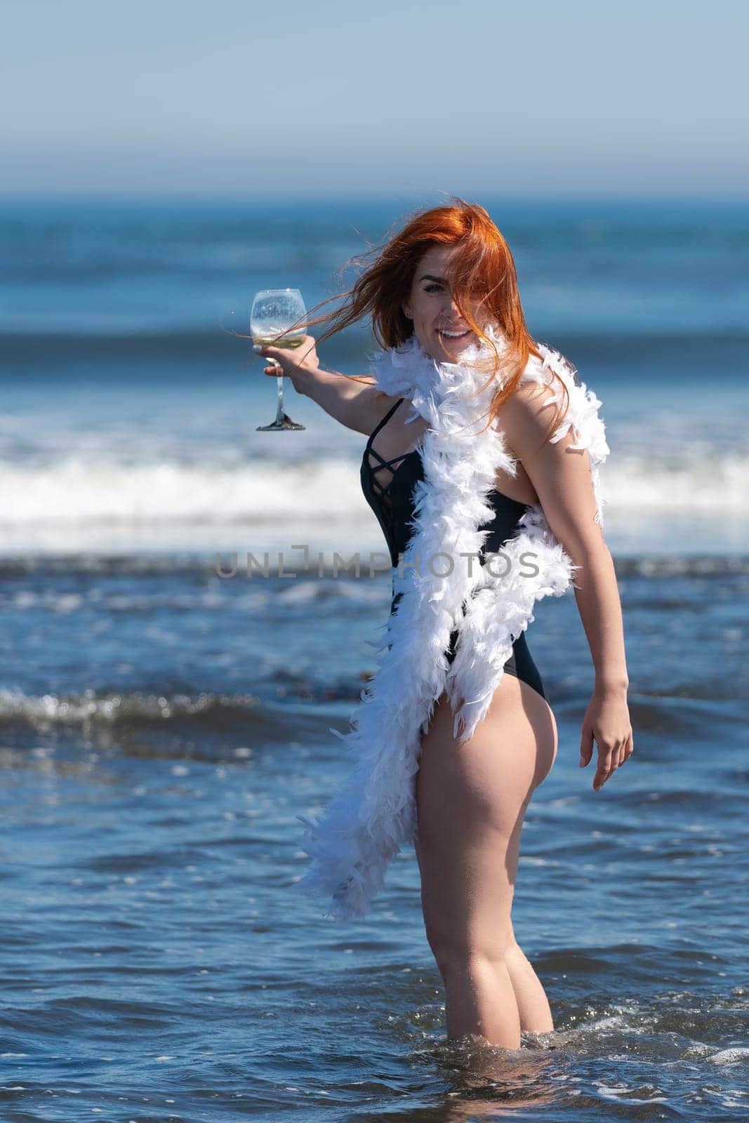 Joyful smiling redhead woman in black one piece swimsuit stands ankle deep in breaking wave of sea and holding glass of wine. Happiness woman has feather boa wrapped around neck. Summer sunny day