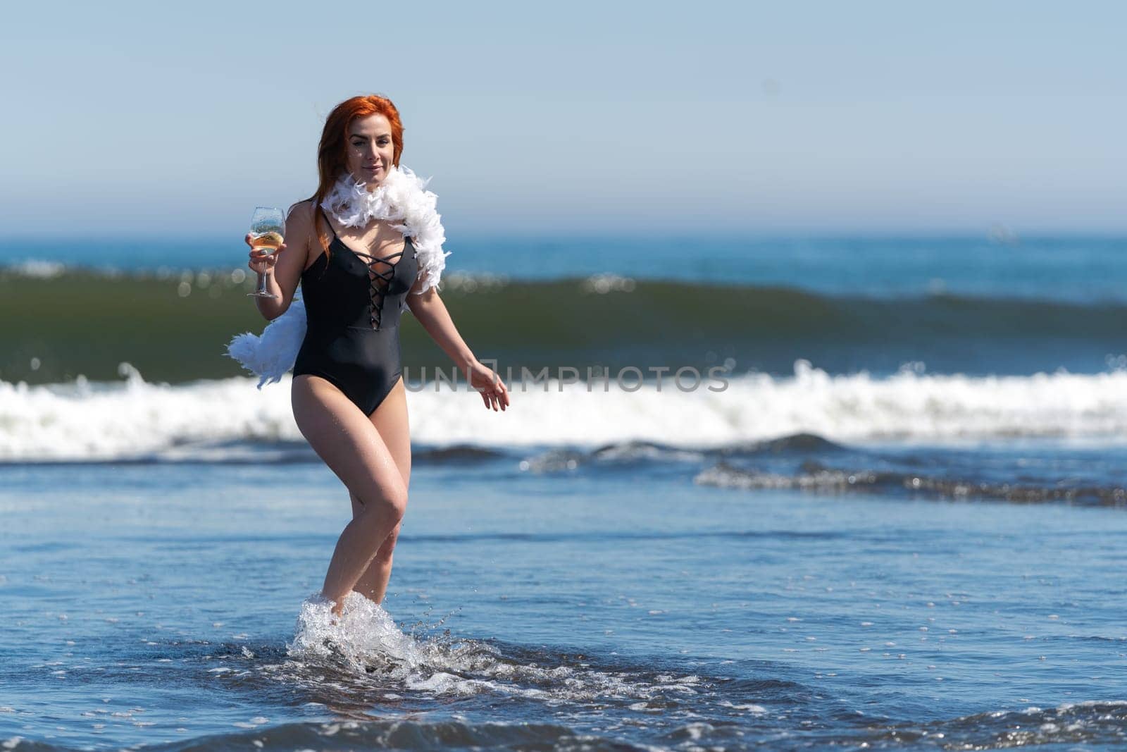 Smiling redhead woman in black swimsuit stands ankle deep in waves of ocean, holding glass of wine by Alexander-Piragis
