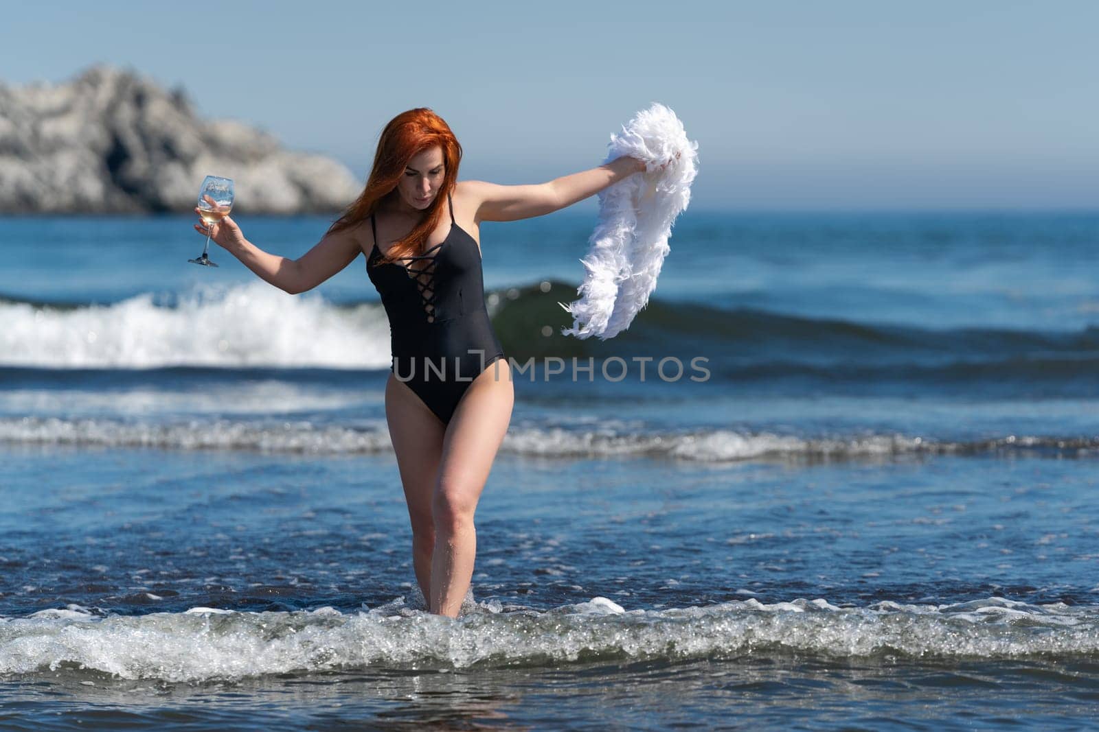 Redhead woman in black one piece swimsuit walks ankle deep into waves of ocean. Female holding wine glass in one hand and boa in other hand. Woman looking down at waves. Front view, full length shot