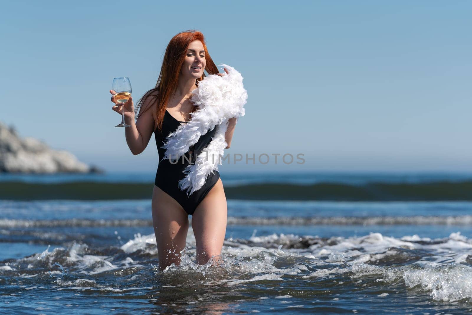 Sensuality redhead woman in black one piece swimsuit walks knee deep in waves of ocean. Female holding wine glass in one hand and boa in other hand. Front view of smiling flirting female on beach