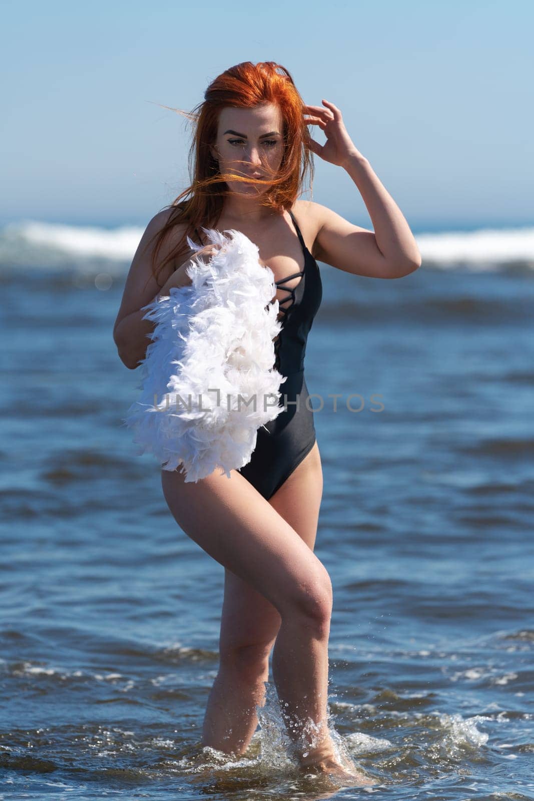 Redhead woman on beach in black one piece swimsuit walks ankle deep into waves of ocean and holds feather boa in hand, straightening her hair in wind with other hand. Woman looking down at sea waves