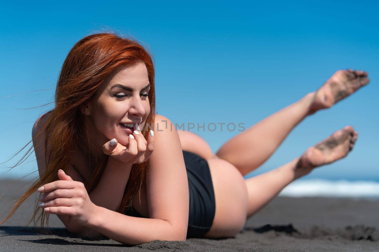 Portrait of happy sunbathing woman on sandy beach with eyes closed. Front view of redhead woman in black one piece swimsuit lying on front with legs raised. Seductive curvy female. Focus on foreground