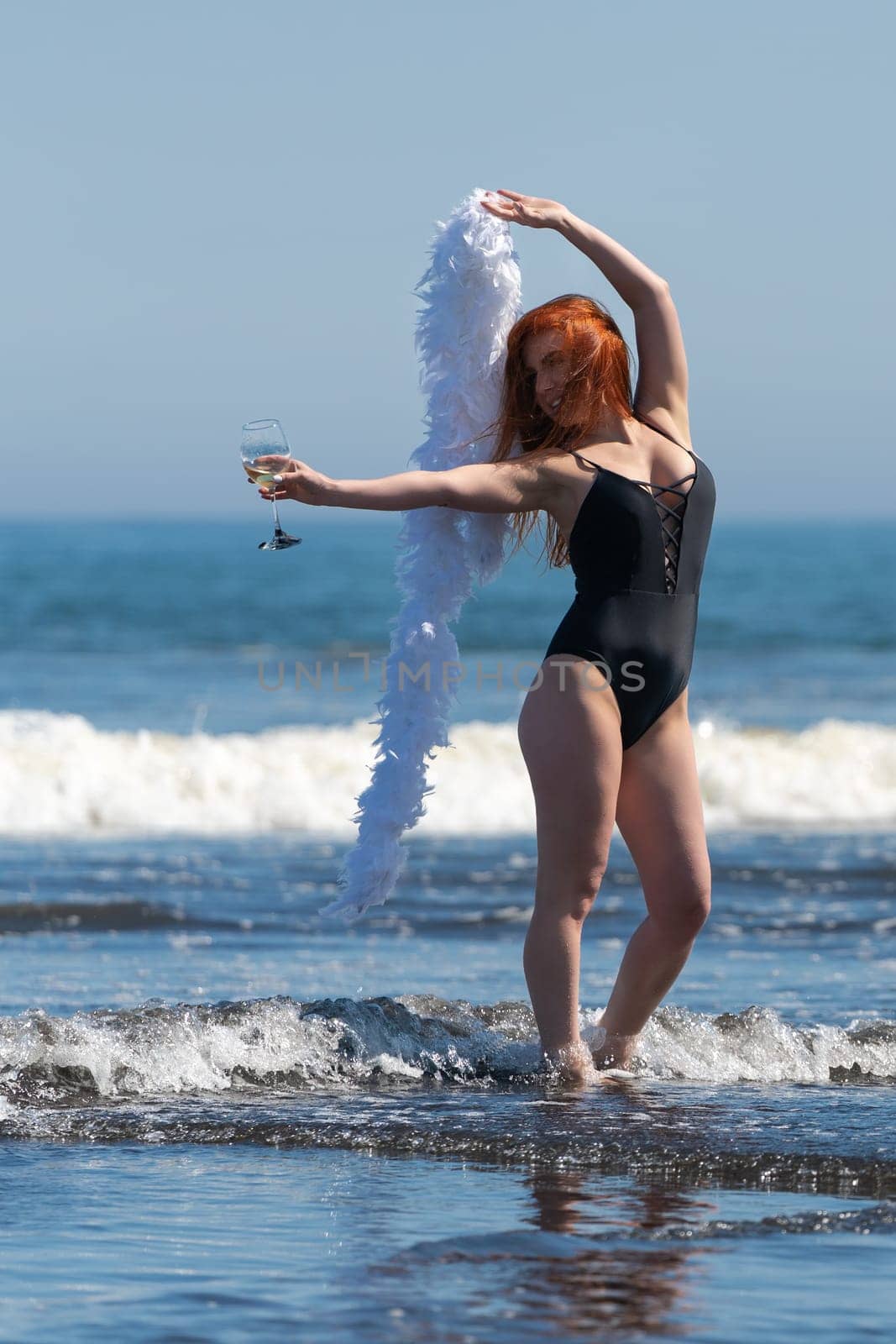 Gorgeous redheaded woman standing ankle deep in ocean waves, holding glass of wine in one hand and boa in other, raised above her head. She looks like having so much fun during summer beach holiday