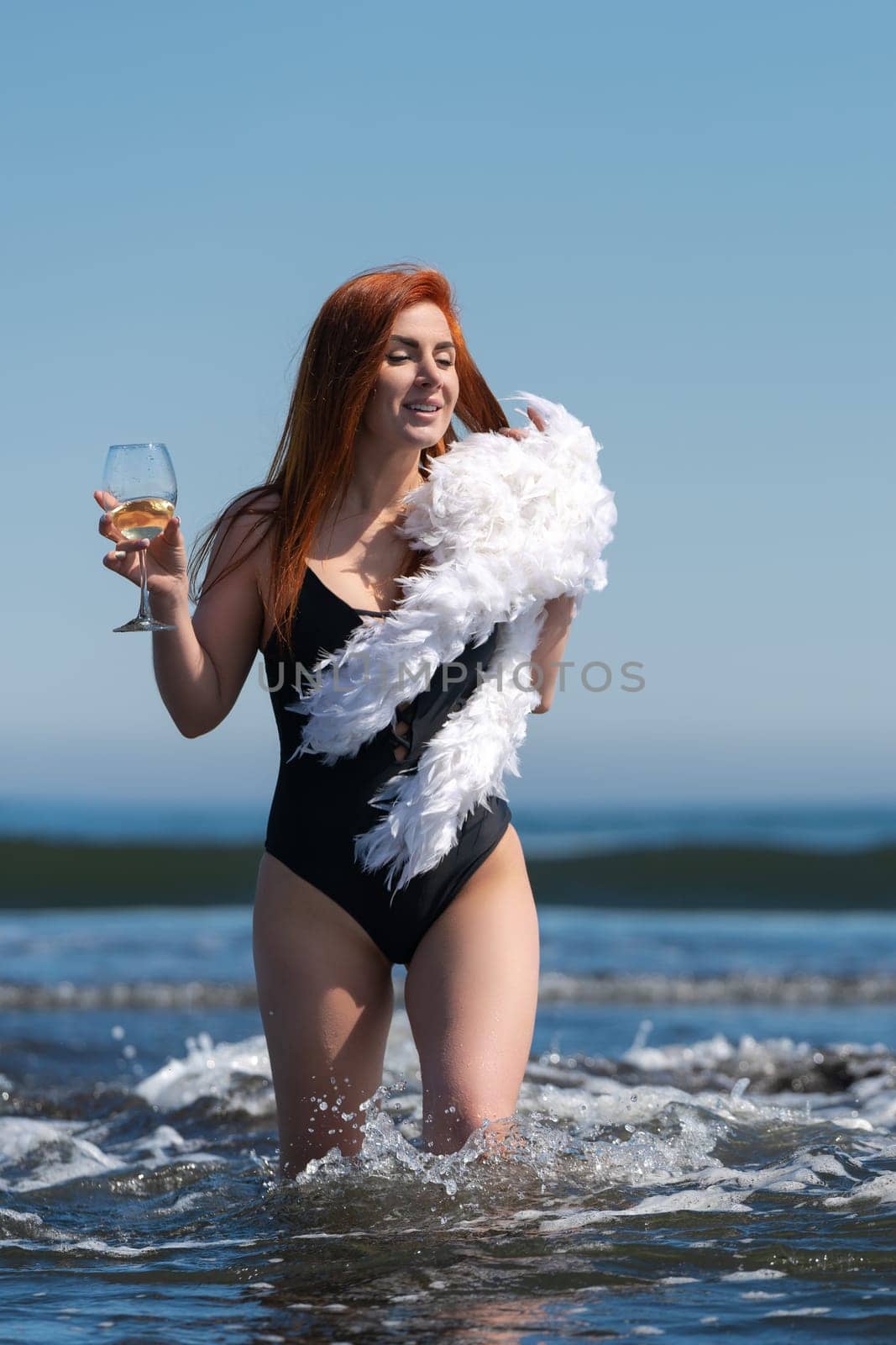 Redhead woman is walking knee deep in waves of ocean, looking fabulous in black one piece swimsuit. And check out that wine glass in one hand and boa in other - she's definitely living her best life