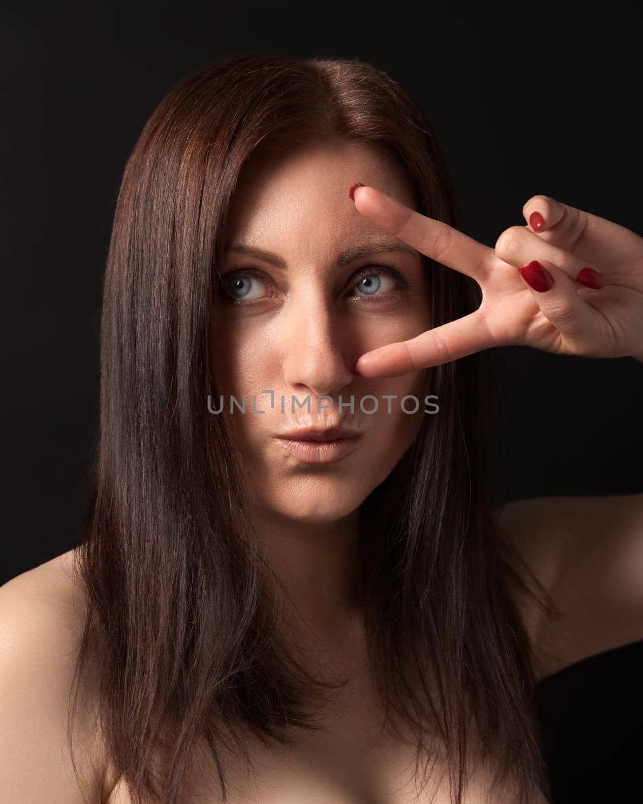 Studio portrait of 40-year-old woman showing victory or peace gesture with fingers near gray eyes. Caucasian ethnicity brunette with long hair and outstretched lips looking away on black background.