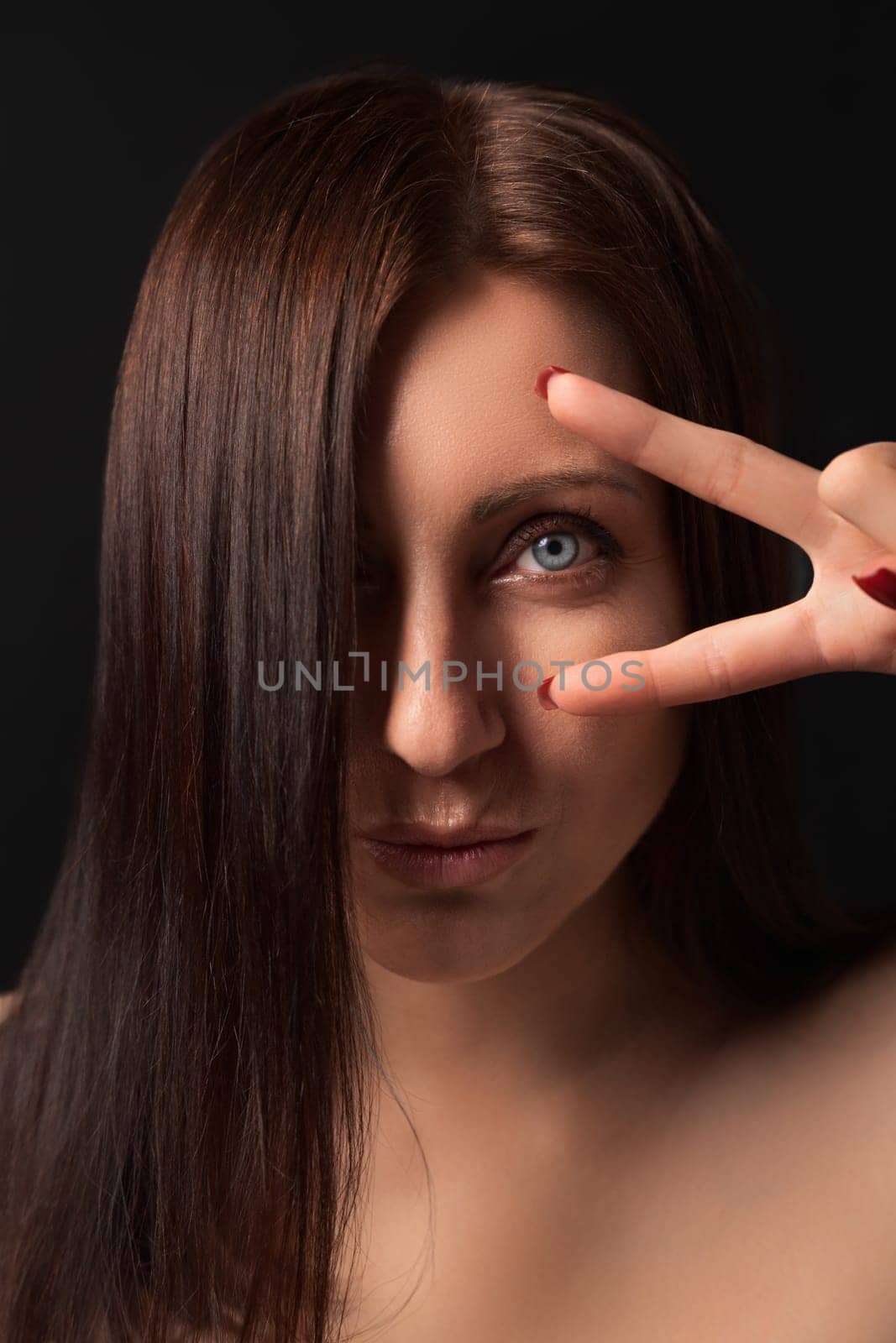 Portrait of nice adult lady showing victory sign near eye and looking at camera. Lovely Caucasian ethnicity brunette woman with long hair covering one eye and outstretched lips on black background.