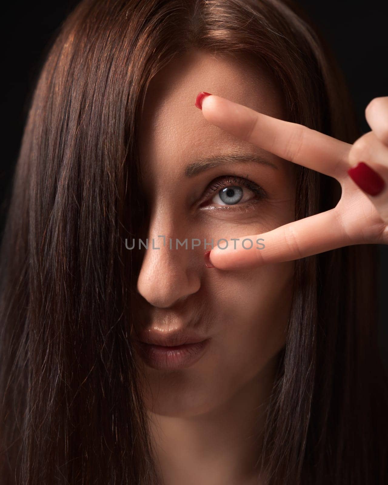 Closeup portrait of woman showing victory or peace gesture with fingers near gray eye and looking at camera. Front view of brunette lady with long hair covering one eye and outstretched lips.