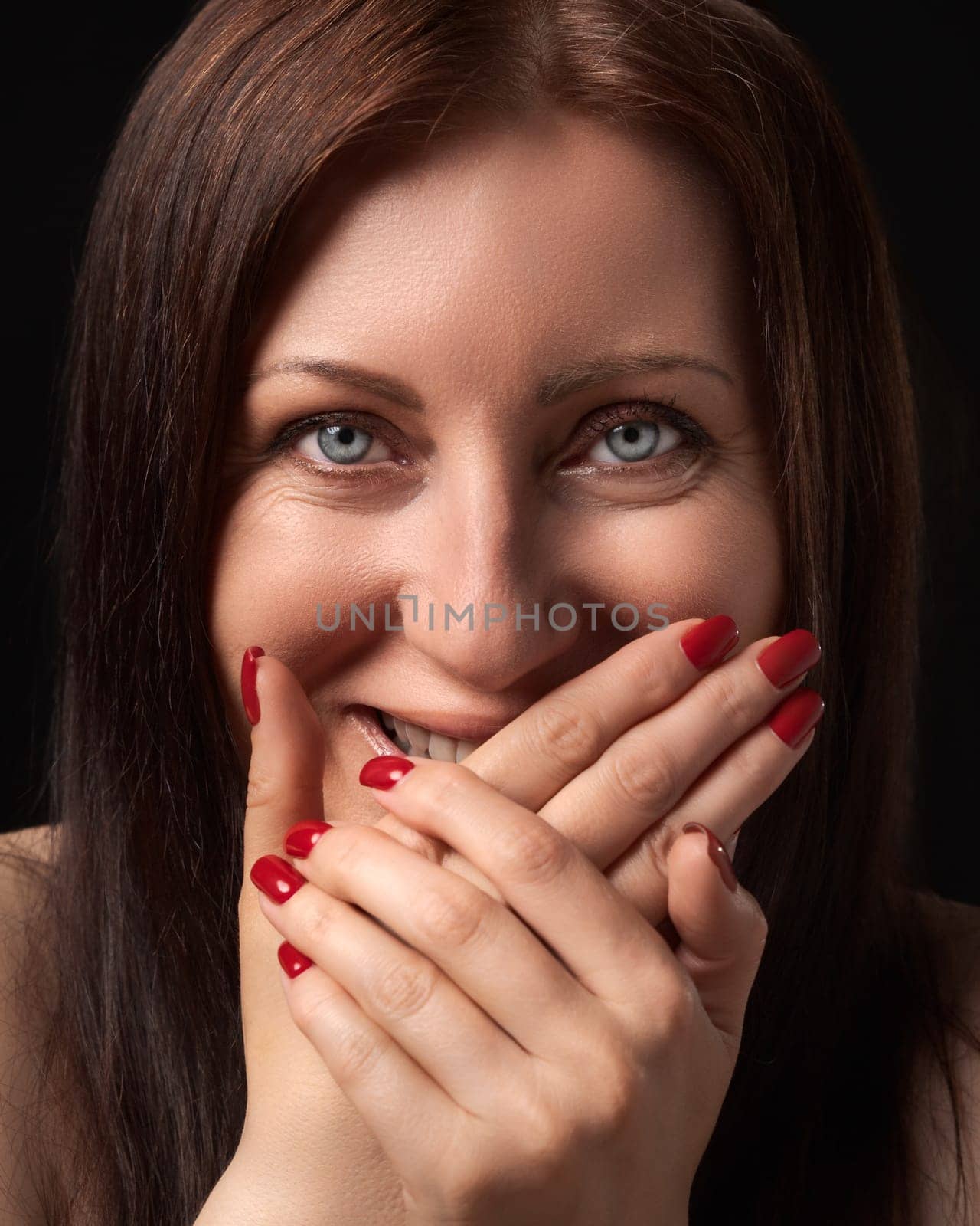 Cheerful charming woman laughing while covering mouth with her hands with red manicure on nails and gazing at camera. Studio portrait of Caucasian ethnicity 40-year-old brunette woman with gray eyes.