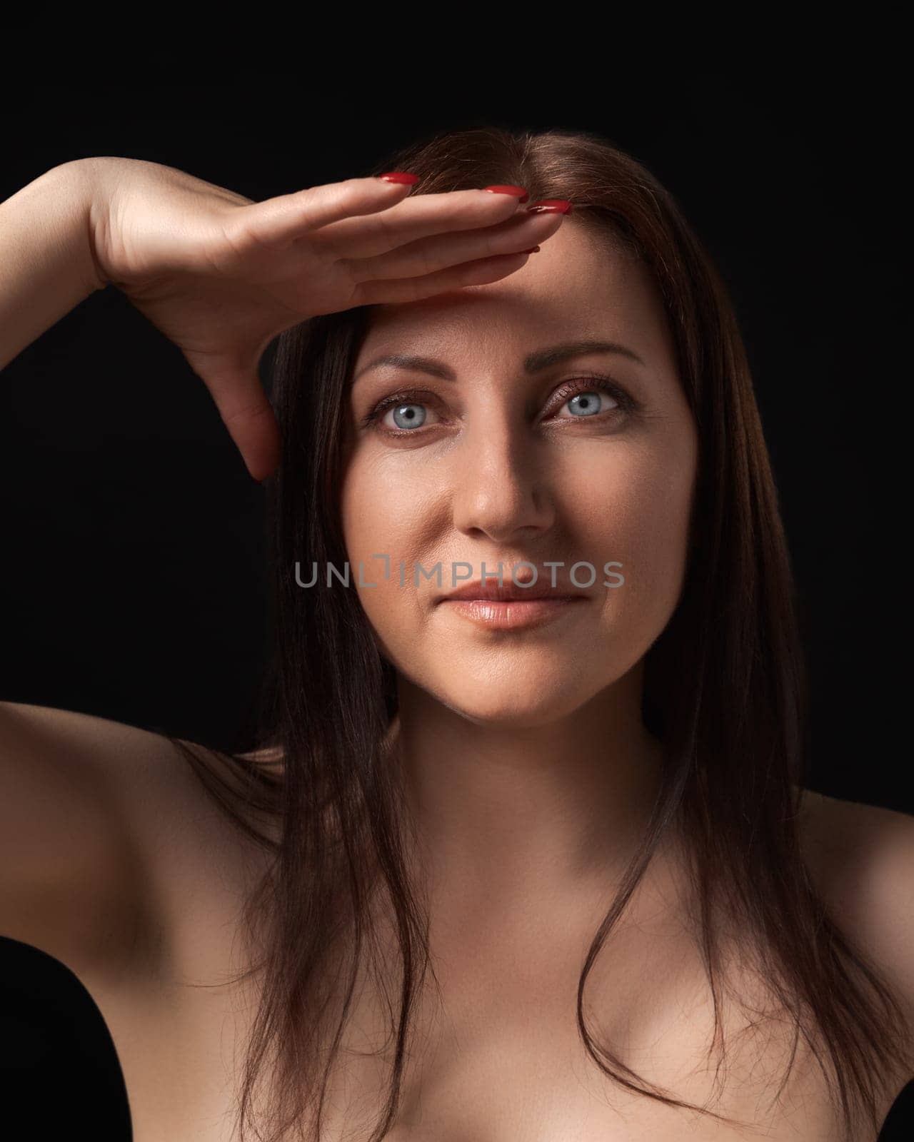 Pretty brunette woman gracefully raising hand to her forehead and looking at camera on black background. Studio portrait of beautiful and sexy woman with gray eyes and long hair. Sincere emotions