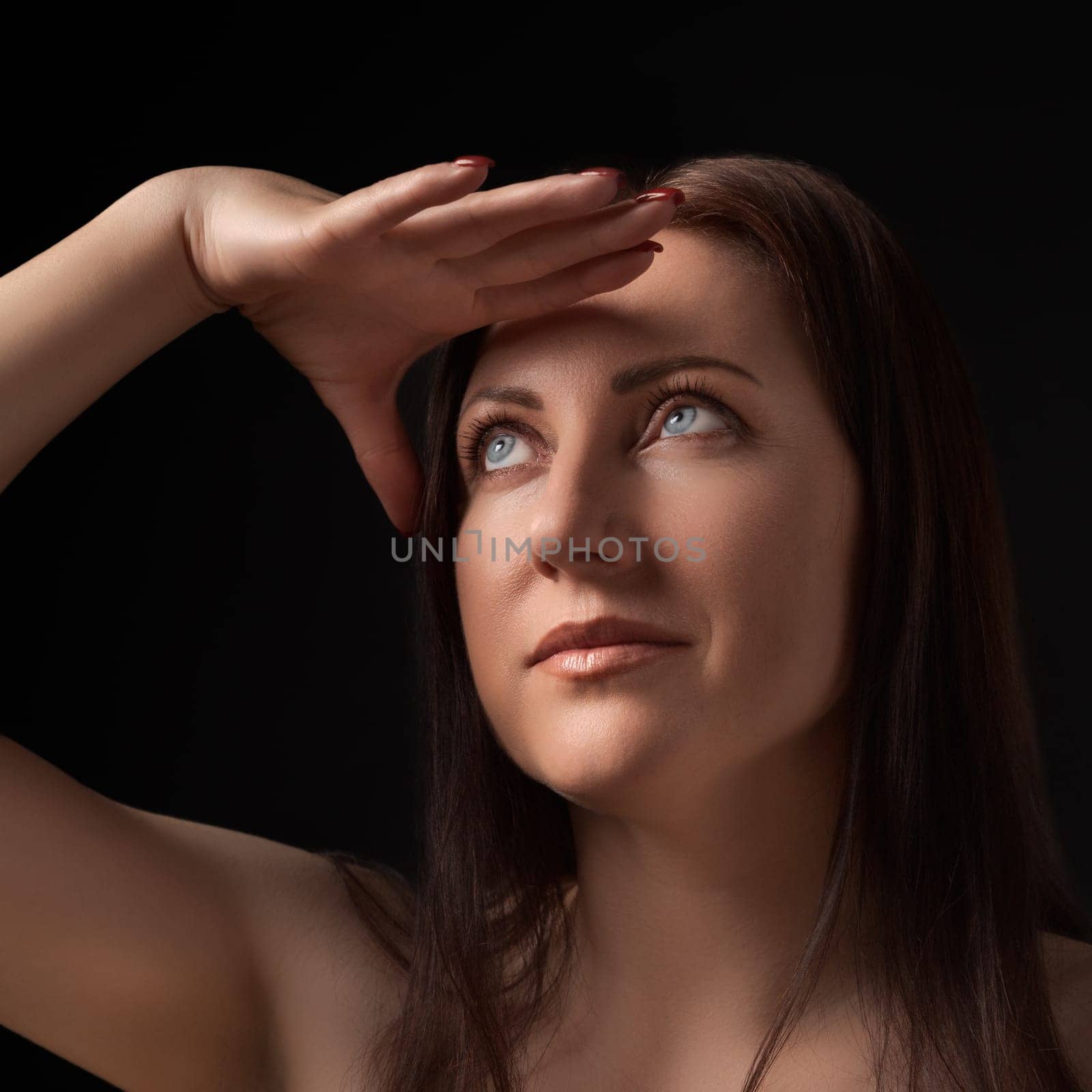 Portrait of beautiful and sexy adult woman proudly raising hand to her forehead and looking up away on black background. Studio headshot of 40-year-old brunette woman with gray eyes and long hair.