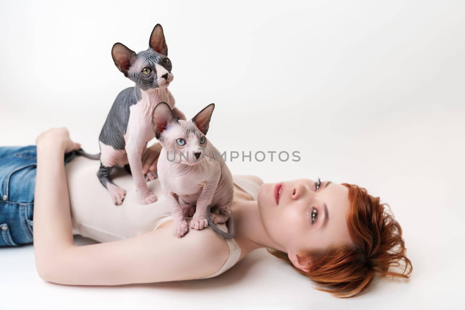 Two friendly Sphynx Hairless Cat, sitting on redhead young woman lying on back on white background. Beautiful woman with short hair wearing T-shirt and blue jeans. Part of series. Selective focus
