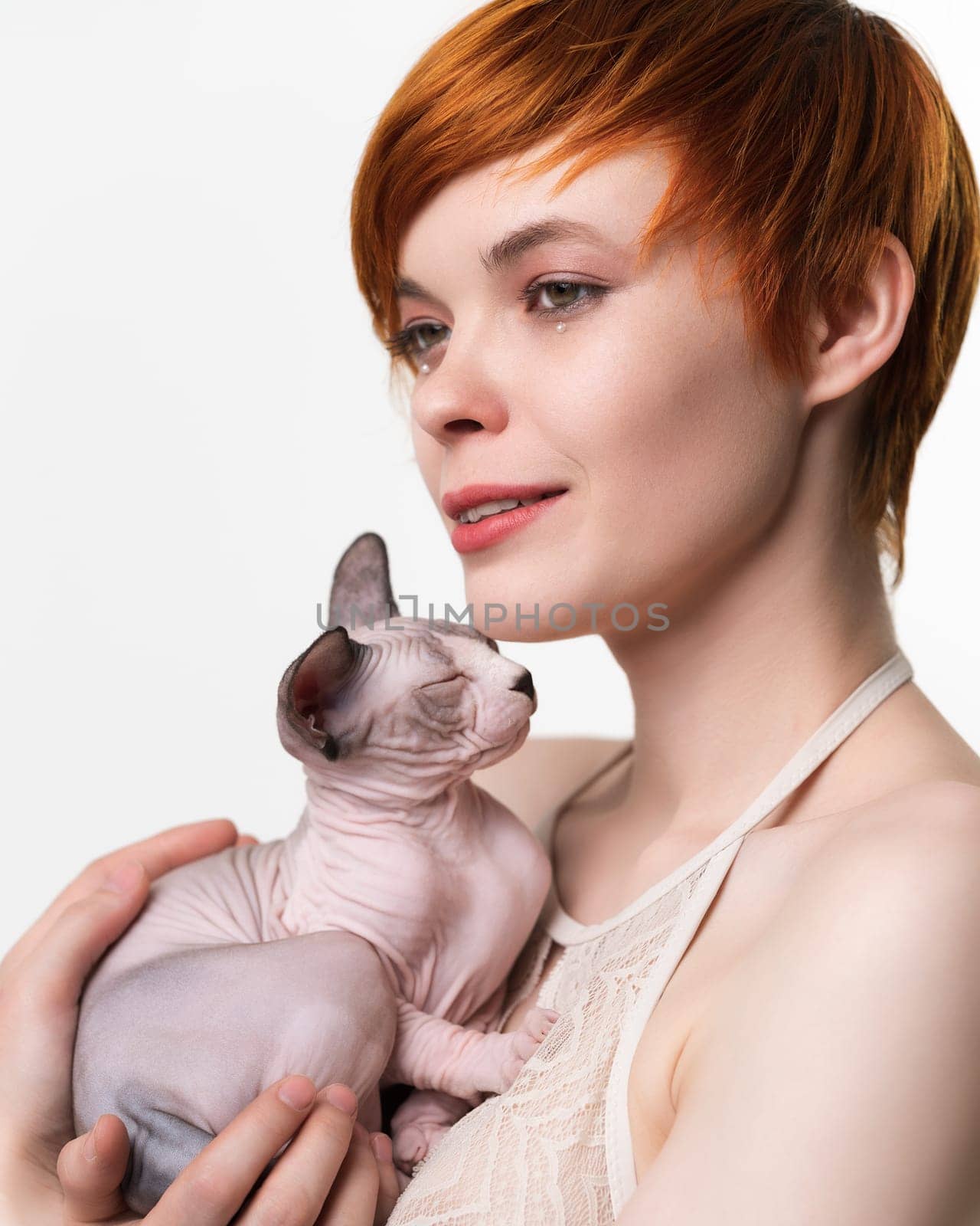 Cute redhead young woman gently hugging sleeping kitten to her chest and looking away. Portrait of pretty woman with short hair 25 years old. Studio shot on white background. Part of series.