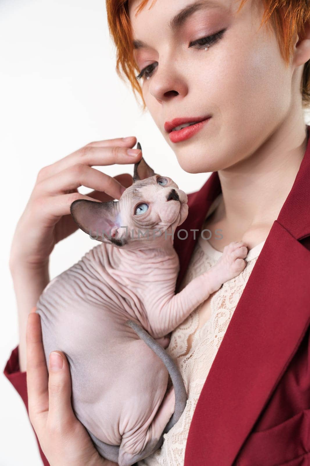 Hipster young redhead woman with short hair gently hugging to her chest Canadian Sphynx Cat. Cute woman looking away, dressed in red jacket. Studio shot on white background. Part of series.