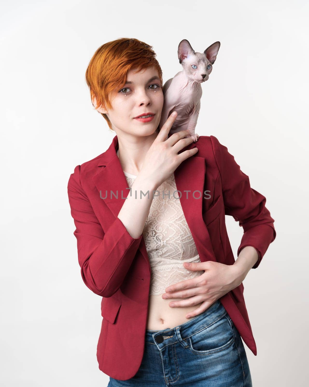 Beautiful hipster redhead young woman looking at camera, dressed in red jacket, blue jeans and playful Sphynx Hairless kitten sitting on her shoulder. Studio shot on white background. Part of series