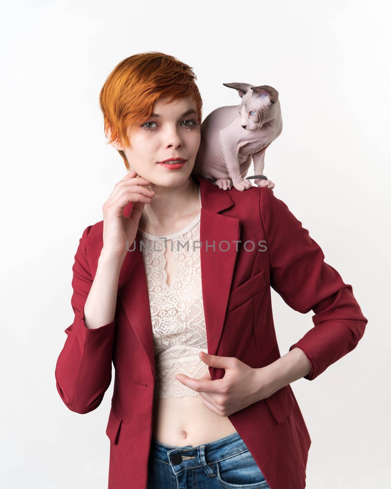 Portrait of hipster redhead young woman with short hair, dressed in red jacket, blue jeans and playful Sphynx Cat sitting on her shoulder. Front view studio shot on white background. Part of series