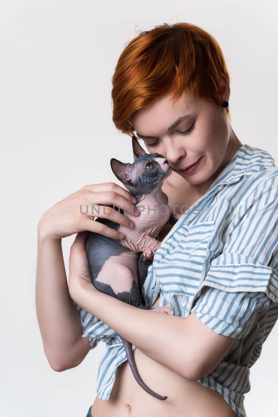 Redhead young woman with closed eyes gently hugs cat looking at owner. Studio shot on white background. Portrait beautiful hipster with short hair dressed in striped white-blue shirt. Part of series.