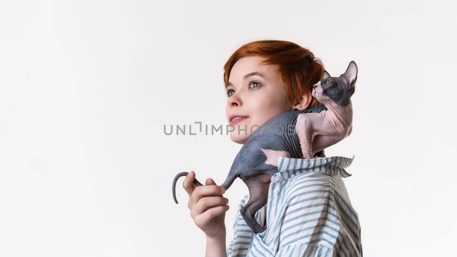 Smiling redhead young woman with Canadian Sphynx Cat sitting on her shoulder. Portrait of hipster dressed in striped white-blue shirt. Studio shot on white background, copy space. Part of series.