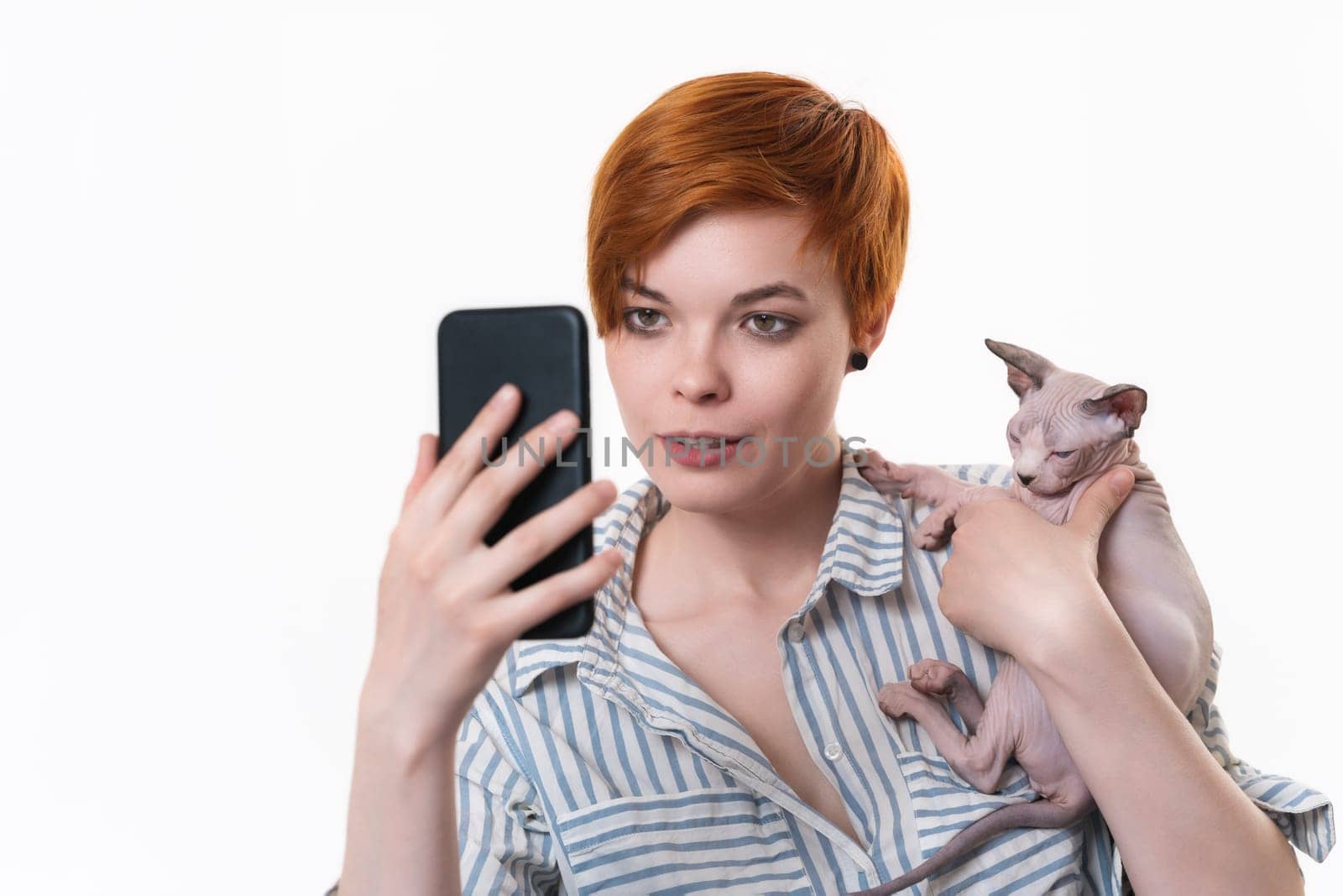 Redhead young woman hugging cat, having video-call holding smart phone in hand shooting selfie on camera. Studio shot on white background. Hipster in striped white-blue shirt. Part of series.