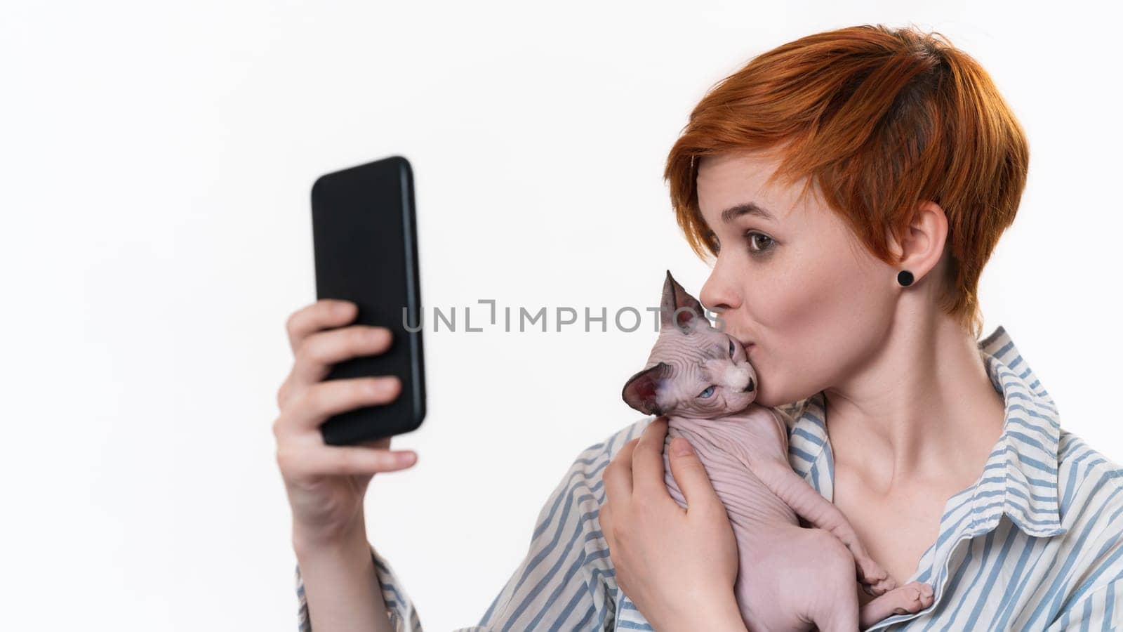 Redhead young woman holding and kissing kitten, taking selfie photo using smartphone camera. Studio shot on white background. Hipster with short hair dressed striped white-blue shirt. Part of series.