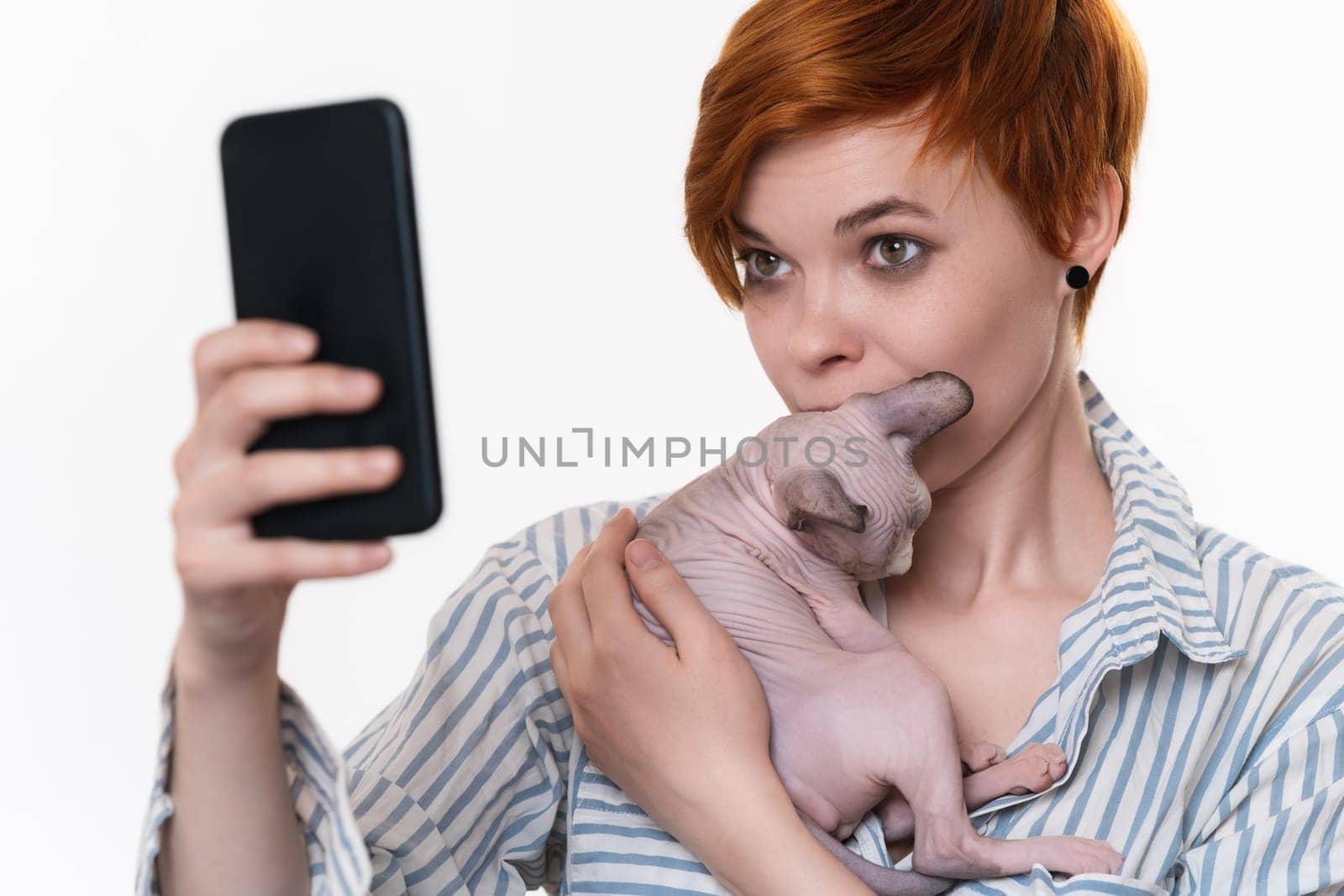 Woman holding and kissing kitten, taking selfie self portrait photos on smartphone. Young adult redhead hipster dressed striped white-blue shirt. Part of series. Studio shot on white background.