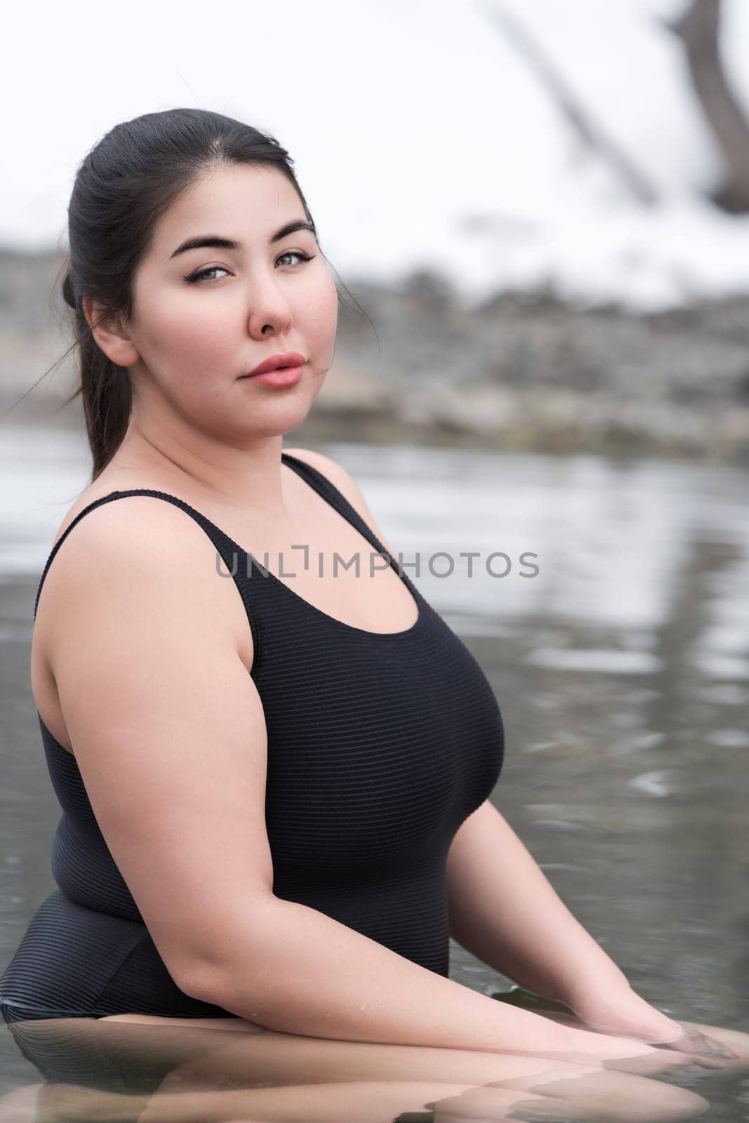 Young busty curvy full figured young adult model in black one piece bathing swimsuit sitting and relaxation in mineral water in outdoors pool at spa resort. Healthy lifestyle, body positive concept
