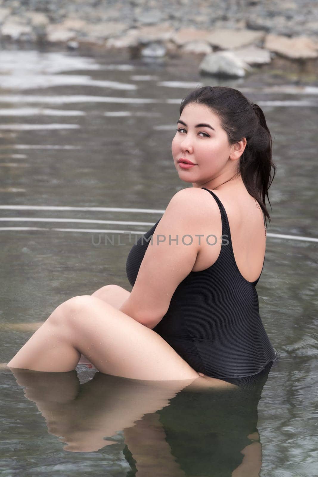 Overweight young woman in black one-piece bathing swimsuit sitting in mineral water in outdoors pool at balneotherapy spa, hot springs resort. Side view of full figured xxl woman looking over shoulder